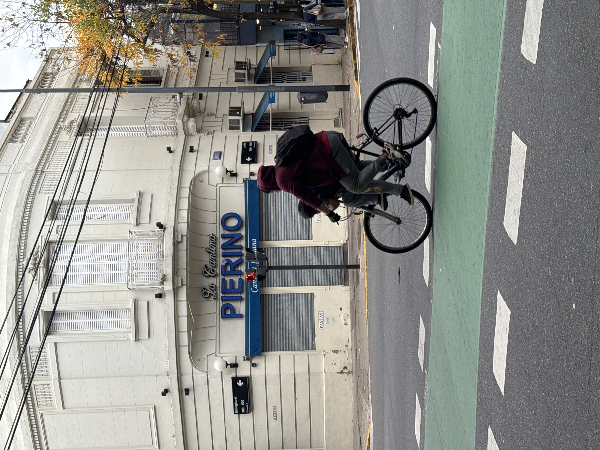 A delivery cyclist rides on a bicycle lane in Buenos Aires, Argentina. The building on the left has a sign reading "La Cantina PIERINO." Street signs indicate Billinghurst 900-800. The day is overcast. The cyclist wears a red hoodie, gray pants, and a backpack. A tree with yellowing leaves is visible. People are standing on the sidewalk.