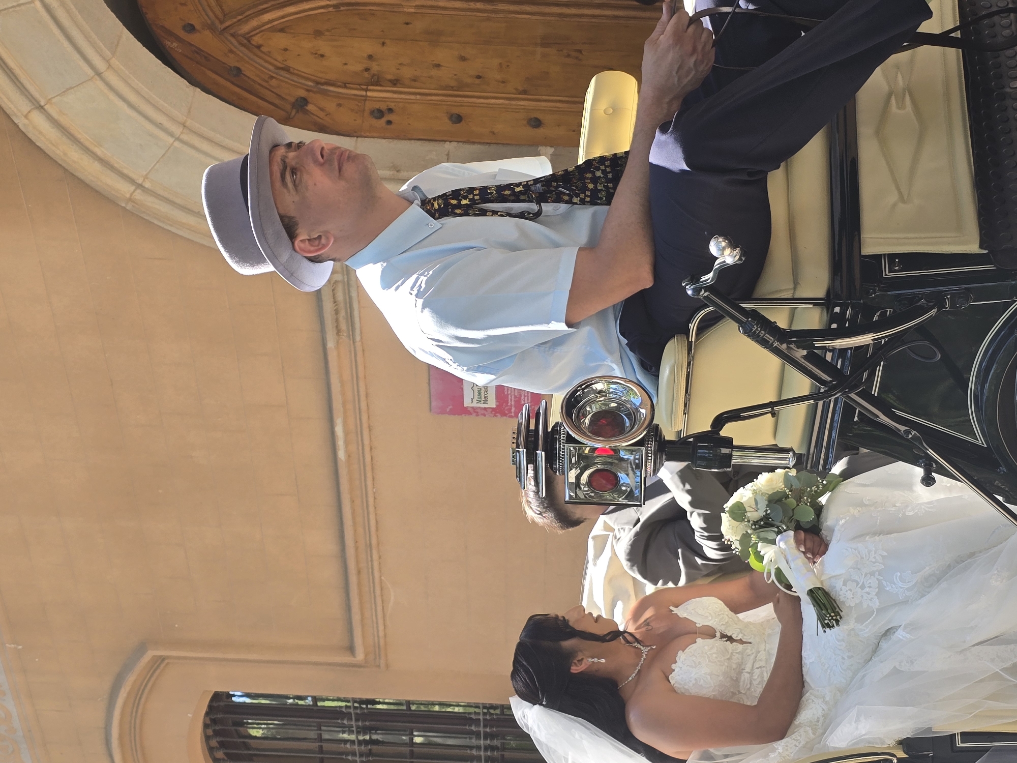 This image depicts a wedding scene, likely in Cornellà de Llobregat, Spain, given the context of previous searches. A bride, adorned in a white lace wedding gown and veil, sits in a horse-drawn carriage holding a bouquet of white roses and greenery. Beside her, a man wearing a light blue short-sleeved collared shirt, a patterned tie, and a grey fedora, also sits in the carriage. He appears to be the carriage driver or a passenger. In the background, a light brown building with arched windows and a wooden door suggests a historic or religious establishment. The carriage itself is ornate, with black metal accents and cream-colored upholstery. A camera-like apparatus with red lenses is mounted on the carriage in front of the passengers. The scene is illuminated by bright sunlight.
