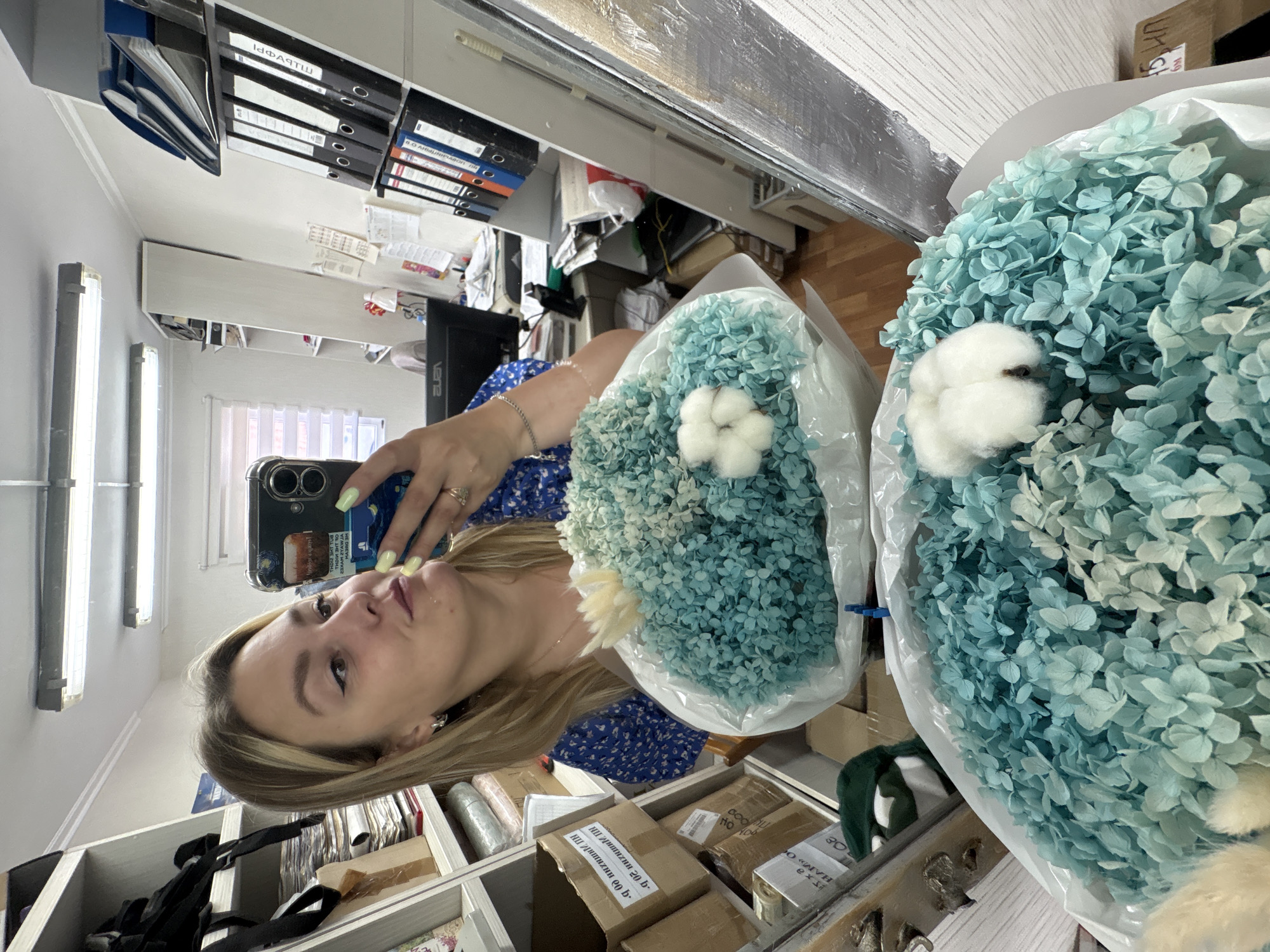 A young woman with blonde hair, wearing a blue floral dress and yellow nail polish, takes a selfie in a room filled with shelves. She holds a smartphone in her left hand and a large bouquet of dried blue hydrangeas with cotton accents in her right. The hydrangeas are wrapped in white plastic. She appears to be in a brightly lit room with fluorescent lights on the ceiling. The shelves behind her are filled with files, boxes, and other office supplies. The room has wooden floors and light-colored walls. Visible text on a box says "Vene".