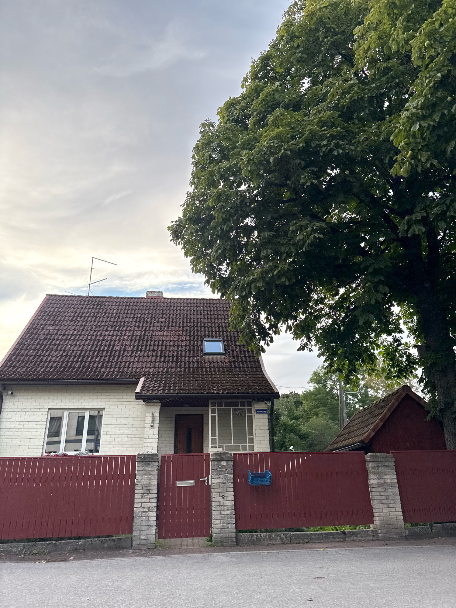 The image depicts a suburban street scene in Tallinn, Estonia. In the foreground, a dark red wooden fence with vertical slats and stone pillars borders a property. A residential house with white brick walls and a dark tiled roof is visible behind the fence. The house has several windows, a prominent entrance with a wooden door, and a skylight on the roof. A large tree with dense green foliage partially obscures the sky and the right side of the house. Adjacent to the main house, a smaller red-roofed building is also visible. The sky is overcast with diffused light. The overall impression is of a quiet residential area.