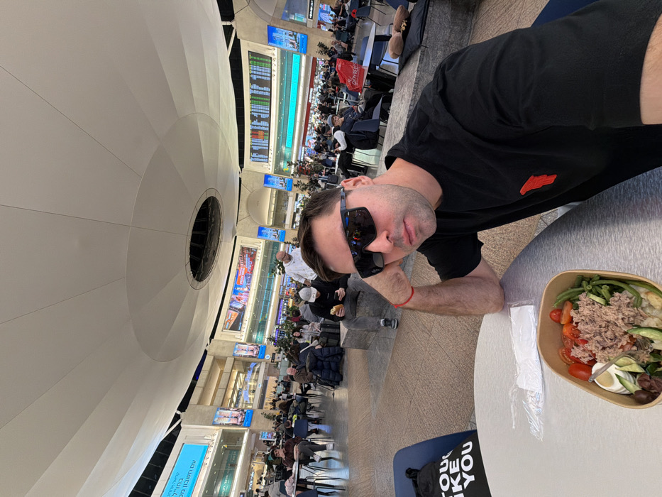 A man wearing sunglasses and a black shirt sits at a table with a salad containing tuna, tomatoes, green beans, and olives. The setting appears to be an airport waiting area, filled with people and large display screens showing flight information. The architecture includes a large, white, dome-shaped ceiling with a central opening.
