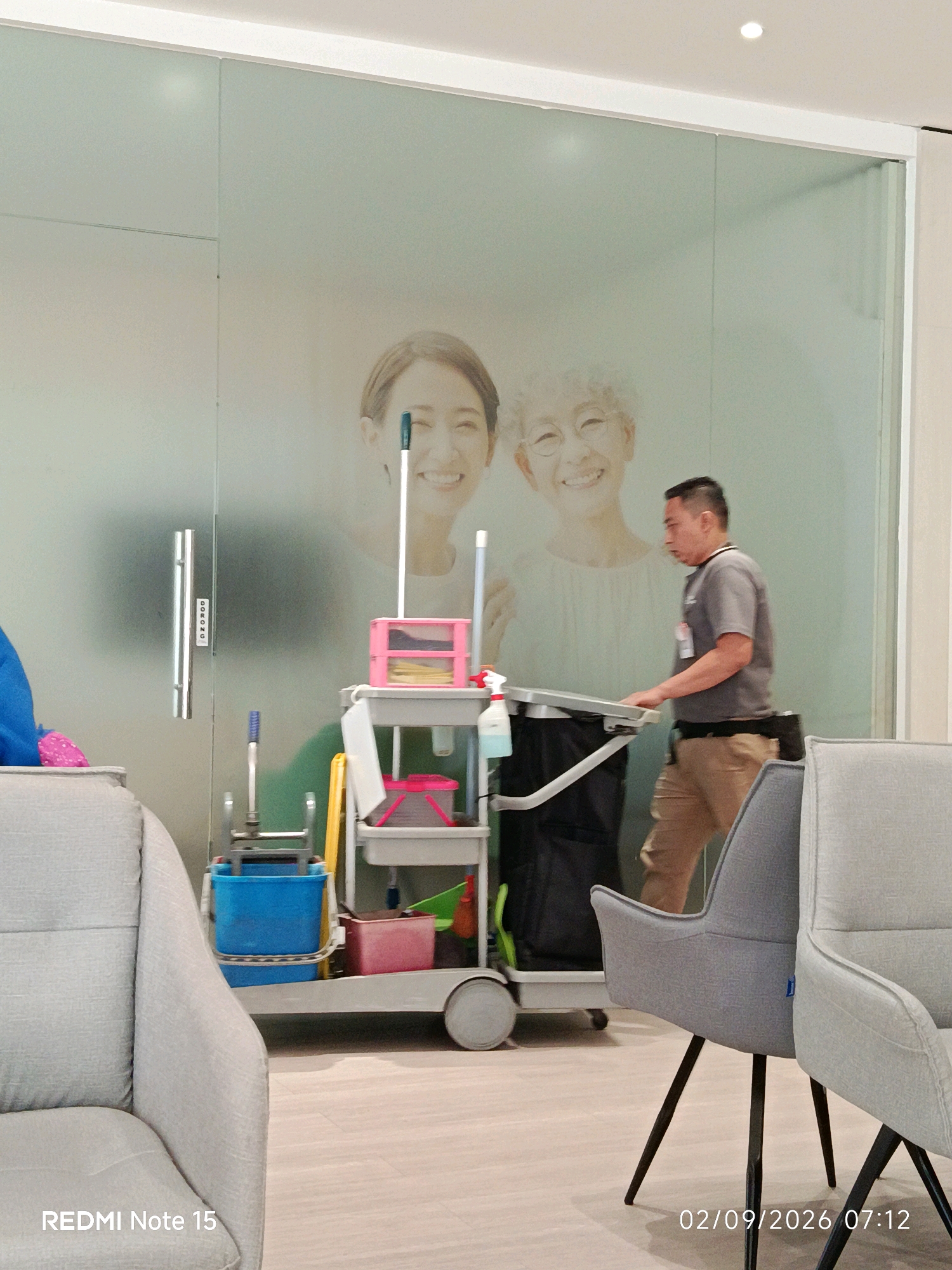 A male janitor is pushing a cleaning cart in front of a frosted glass wall. The wall has a large, faint image of two smiling women on it. The janitor is wearing a grey t-shirt and tan pants and is looking down at the cart. The cart contains cleaning supplies such as buckets, mops, and bottles. In the foreground, there are two grey chairs. The floor is light-colored wood. The text "REDMI Note 15" is in the bottom left corner, and "02/09/2026 07:12" is in the bottom right corner. The scene appears to be in a clean, indoor environment, possibly a waiting room or office. The location is Waru, Indonesia.