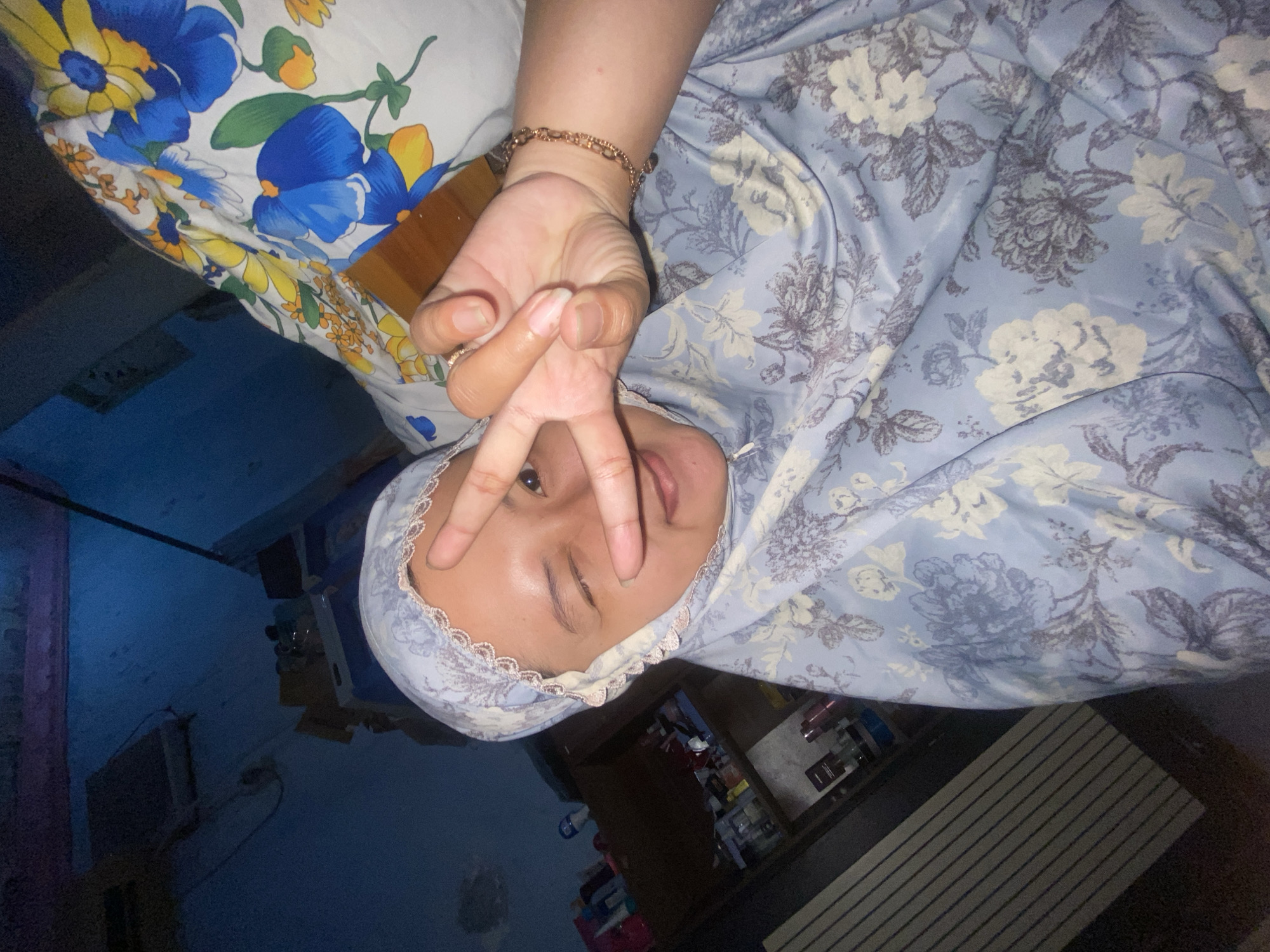 A medium shot shows a young woman from the chest up, posing for a selfie in what appears to be a dimly lit room. She is wearing a light blue hijab with a floral pattern and a white, floral-patterned sheet is draped over her shoulder. She is winking with her right eye and holding up two fingers in a peace sign with her right hand. Her left eye is open. A bracelet made of gold-colored links is visible on her right wrist. In the background, a shelf unit is visible, filled with various bottles and containers. A striped object, possibly a mattress or furniture, is also visible on the left side of the frame. The walls of the room are a dark blue color. The overall lighting suggests it is nighttime. The image was taken in Gelam, Indonesia.