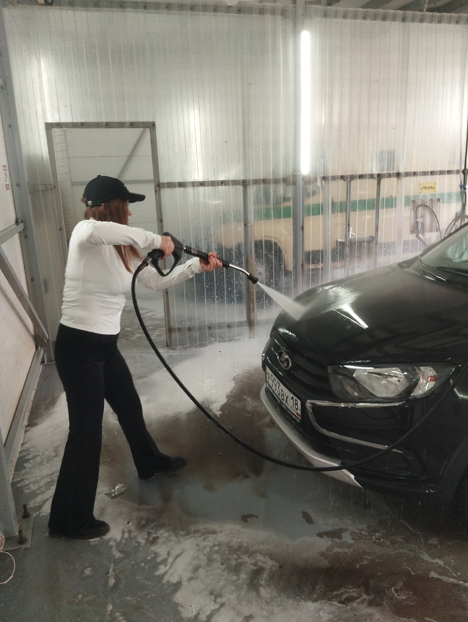 A woman is washing a black car in a car wash bay. She is wearing a white long-sleeved shirt, black pants, black shoes, and a black baseball cap. She is holding a black pressure washer and spraying water onto the hood of the car. The car has a license plate that reads "P993 BX 18."
The car wash bay has translucent corrugated walls and a concrete floor covered in water and soap. A metal frame is visible in the background. The location is in Votkinsk, Russia.