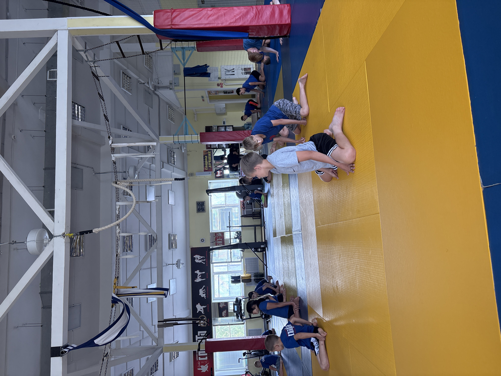 In the city of Chaikovsky, Russia, a group of children are engaged in a judo or martial arts training session inside a gymnasium. The room features a light-colored ceiling with exposed beams and fluorescent lighting. The floor is covered with a tatami mat, predominantly yellow with blue sections. Several children, dressed in blue t-shirts and dark shorts, are positioned on the mat, some sitting with their legs extended, others in a kneeling or crawling posture. In the foreground, a boy in a gray t-shirt and black shorts is seated on the yellow mat. The background reveals gym equipment, including weight racks, and posters on the walls, one with figures demonstrating martial arts poses. Red padded posts are visible along the perimeter of the matting area. The overall atmosphere suggests an active training environment.