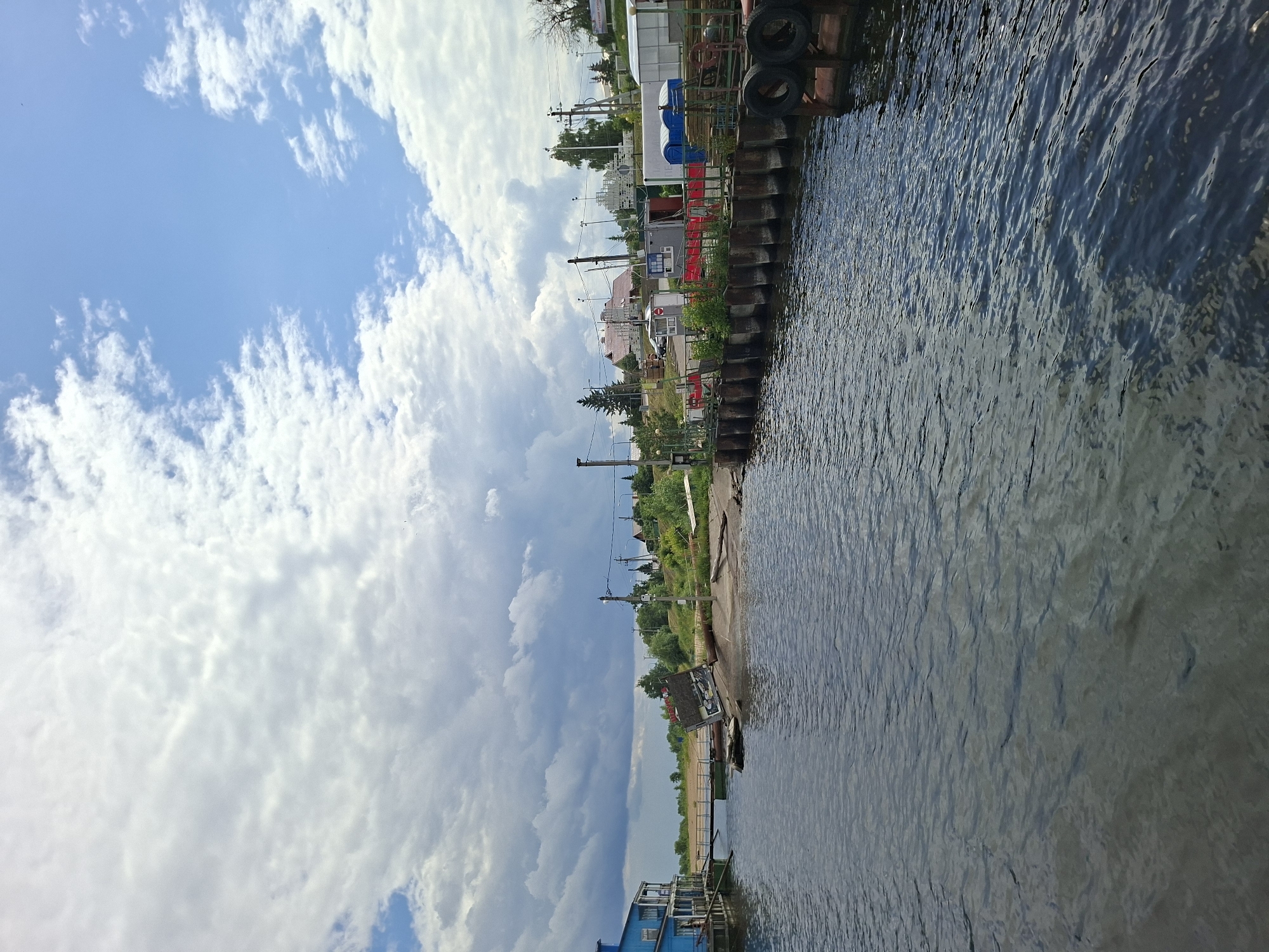 The image depicts a waterfront scene in Verhnij Uslon, Russia. The perspective is from a slightly elevated position, looking out over a body of water that appears to be a river or lake. The water is dark blue and shows ripples, indicating some movement. Along the right edge of the image, a wooden pier or dock is visible, constructed with large wooden beams and reinforced with tires.
On the left side of the frame, the shoreline is lined with various structures. These include what appear to be small buildings, possibly shops or residences, with colorful facades and pitched roofs. There are also utility poles with wires running between them, suggesting the presence of electricity and communication lines. Beyond the immediate buildings, a gentle slope leads up to more trees and vegetation.
The sky is a prominent feature, filled with a mix of bright blue sky and large, dramatic white and grey clouds. The cloud formations suggest a dynamic weather pattern, perhaps a transition from sunny to potentially stormy conditions.
The time of day appears to be daytime, with ample natural light illuminating the scene. The overall mood is calm and somewhat rustic, suggesting a relatively quiet, possibly rural or suburban, area. No people are clearly visible in the image. The scene is characteristic of a riverside settlement in Russia.