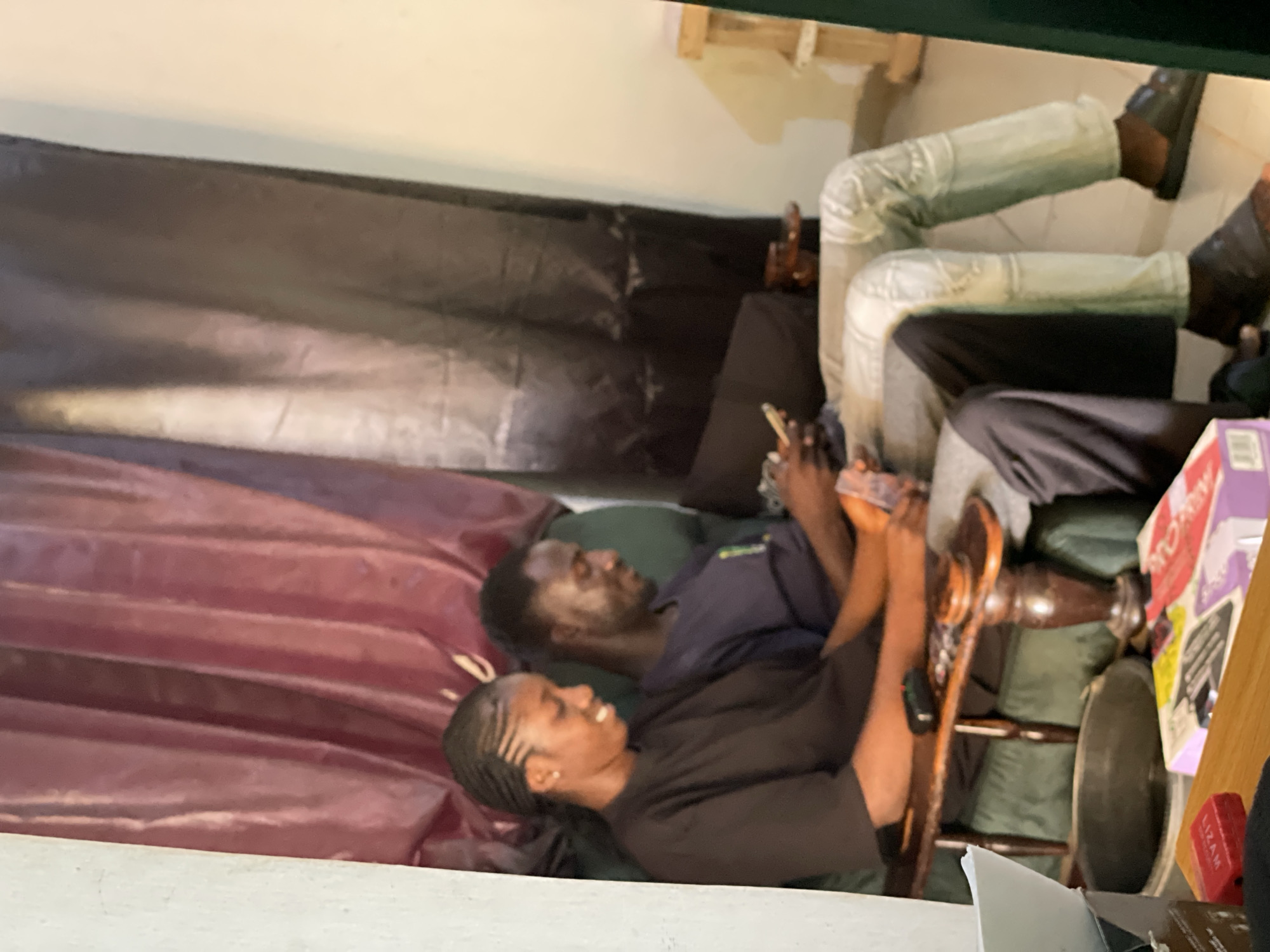 In Durumi, Nigeria, a young man and woman are seated on a green sofa. The woman, on the left, has her hair braided and is wearing a black t-shirt. She is smiling and looking down at a mobile phone in her hands. The man, on the right, is wearing a dark blue t-shirt and light-colored, distressed jeans. He is also looking down at a mobile phone. Between them, a wooden side table holds a remote control and a small circular object. In the foreground, a table displays several items, including a red box labeled "LIZAM" and a box with colorful graphics. The background features dark curtains with a reddish hue.