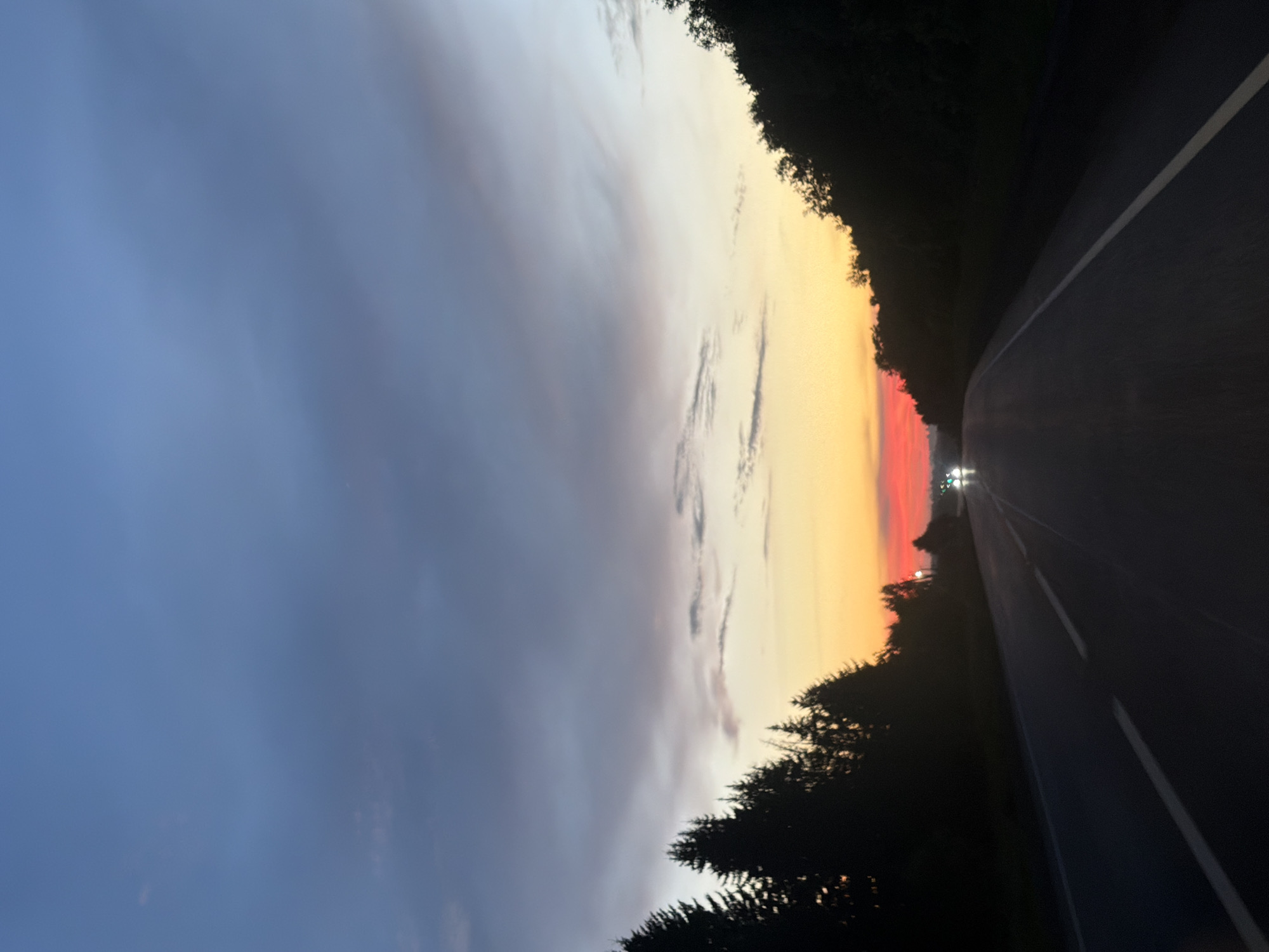 The image depicts a road at dusk, with a gradient of sky colors ranging from deep blue at the top to orange and red near the horizon. Silhouetted trees line both sides of the road, creating a natural border. In the distance, headlights from two vehicles are visible, suggesting traffic on the road. The scene is devoid of any discernible buildings or structures, suggesting a rural or natural setting. The location in the city of Bol'shaya Ven'ya, Russia is not identifiable from the visual information provided in the image.
