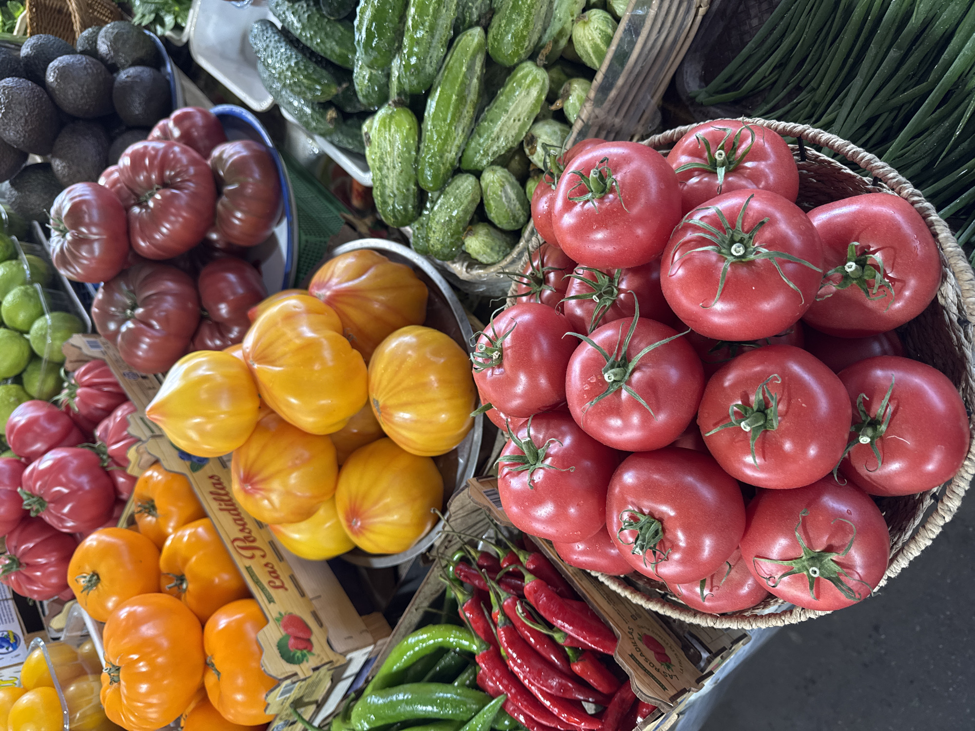 A variety of fresh produce is displayed at a market. In the foreground, a basket overflows with vibrant red tomatoes. To the left, another basket holds bright red chili peppers and green peppers. Behind them, orange and yellow heirloom tomatoes are arranged in wooden crates. Further back, piles of dark green cucumbers and several dark-skinned avocados are visible, along with some green limes. The scene suggests a bustling outdoor market in Odesa, Ukraine, with an emphasis on fresh, colorful ingredients.