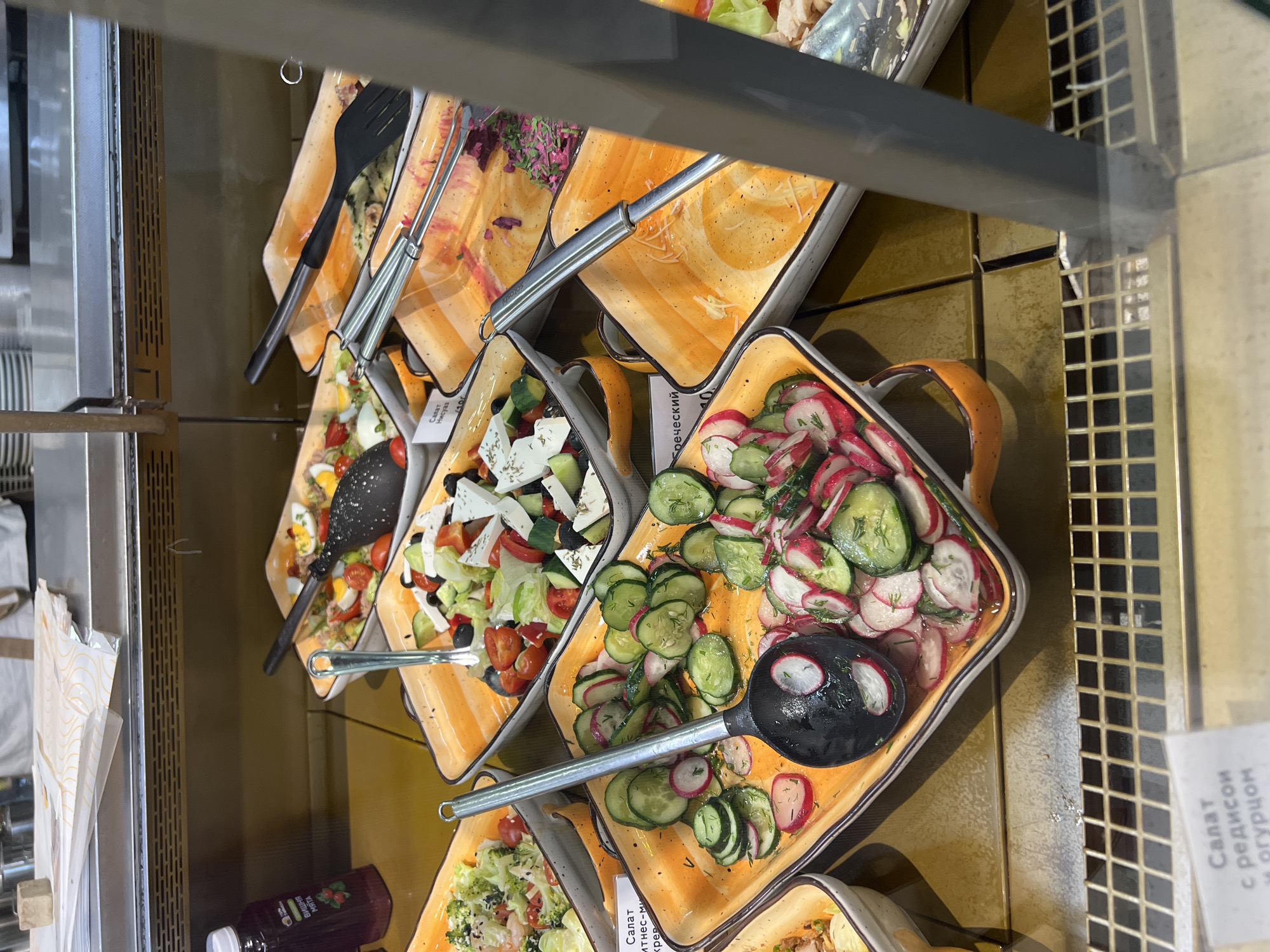 In Yekaterinburg, Russia, at a food service establishment, several salads are displayed in orange rectangular trays within a refrigerated case. In the foreground, a salad of sliced cucumbers and radishes with dill is prominently featured, with a black serving spoon resting in it. To the left, another tray holds a salad with what appears to be cauliflower and possibly other vegetables in a creamy dressing. Behind these, a Greek salad is visible, containing feta cheese, tomatoes, cucumbers, olives, and lettuce, served with a metal tongs. Further back, a salad with sliced hard-boiled eggs, tomatoes, and lettuce is present. The refrigerated case itself has a metallic grid on the bottom, and a bottle of purple liquid is seen to the far left. A white label with Cyrillic text is visible on the bottom left, partially obscured by the grid.