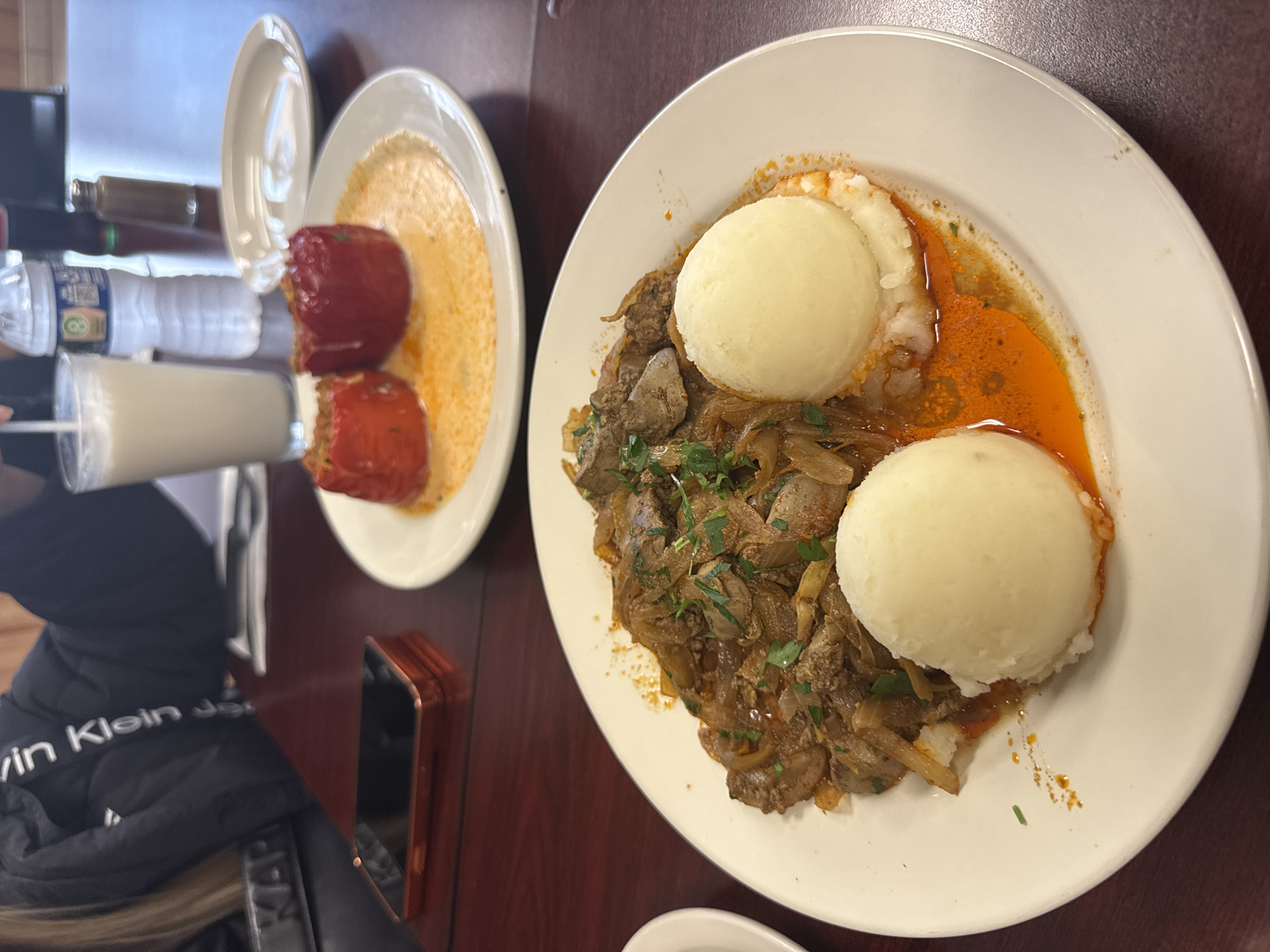 The image shows a table setting in a restaurant, possibly in Des Plaines, United States of America. Two plates of food are visible. The plate on the left contains what appears to be stuffed peppers in a creamy sauce. The plate on the right features two scoops of mashed potatoes, liver and onions, and a reddish-orange sauce or gravy. A glass of milk, a water bottle, and a bottle of condiment are situated next to the plate of stuffed peppers. A Calvin Klein jacket and a cell phone are near the plates. The table has a dark brown surface.