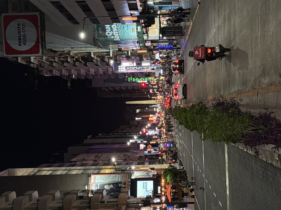 The image captures a nighttime urban scene along Avenida Corrientes in Buenos Aires, Argentina, looking southeast towards the Obelisco, which is visible as a prominent white monument in the distant center.
The street is a multi-lane asphalt thoroughfare, with vehicular traffic currently stopped at a red light. In the immediate foreground, a person on a red scooter, wearing a helmet and a dark jacket, is positioned in the rightmost lane, facing away from the viewer. A red "PedidosYa" delivery box is mounted on the rear of the scooter, which bears a license plate with the characters 'AA 186HA'. Further down the street, a white compact car and a dark sedan are stationary in the same lane. The street features a central median strip planted with green shrubs and smaller purple foliage.
Tall, multi-story buildings line both sides of the avenue, characterized by numerous balconies and a dense array of commercial signage. On the left side, a large vertical billboard displays "MUJER ARAÑA" with an image of a bald man, and another sign below reads "GERMAN KRAUS." An outdoor cafe area with red umbrellas and patrons is visible on this side. On the right, a prominent red billboard with a white circle advertises "PUBLICITE 4556-1701," while a green billboard below it displays "¿Qué somos?".
Pedestrians are visible on both sidewalks, walking and standing. On the left side, a person in yellow clothing is seen near the curb with a small dog. The scene is illuminated by numerous streetlights, bright commercial signs, and active red traffic signals, indicating a busy urban environment after dusk.