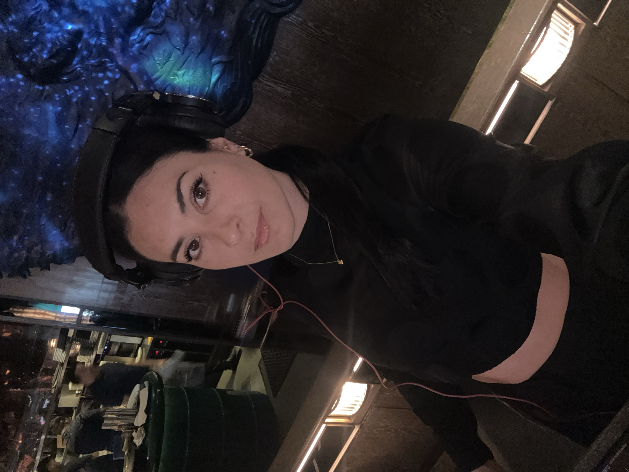 A medium indoor shot shows a woman with black hair, wearing black headphones, a black top, and a gold necklace. She is positioned slightly off-center to the left, looking directly at the camera with a slight smile. A thin pink wire is visible, connected to her headphones. Behind her to the left, the out-of-focus cityscape suggests a high-rise building. To the right, a decorative, dark-colored, possibly wooden or metal structure is visible, lit with blue light. To the right, there is also a wall with illuminated vertical bars. The environment implies an upscale venue, potentially a restaurant or lounge. While the image context suggests London, there are no definitive markers in the frame to confirm that location.