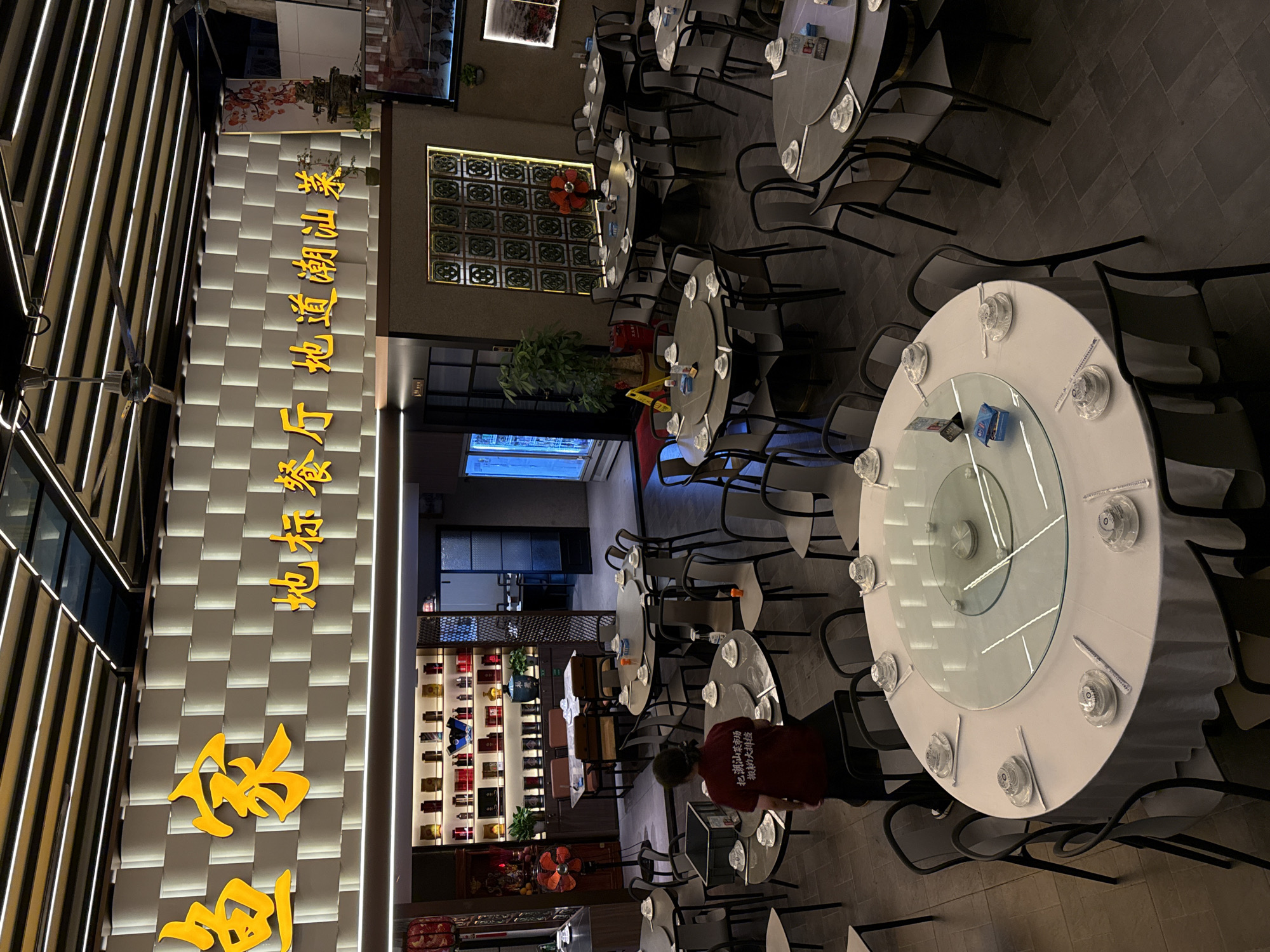 This image captures the interior of a Chinese restaurant, likely in the Nanshan District of China, featuring a spacious dining area with multiple round tables set for customers. Yellow Chinese characters, translating to "Fisherman's Home, Authentic Chaoshan Cuisine," are prominently displayed on a textured wall above the dining space. The restaurant has a modern design with overhead lighting and ceiling fans.
In the foreground, a woman in a red shirt with Chinese writing on the back is seen standing near a round table. The tables are covered with white tablecloths and set with individual place settings, including bowls, chopsticks, and napkins. Some tables have lazy Susans in the center. The dining area is well-lit, with a mix of ambient and direct lighting.
Behind the main dining area, there's a visible counter or display shelf filled with bottles and jars, possibly containing drinks or ingredients. Further back, a doorway leads to another section of the restaurant, with additional tables visible. Decorative elements, such as a bonsai tree and artwork on the walls, add to the ambiance. The flooring appears to be tiled. The overall impression is of a clean and inviting dining establishment.