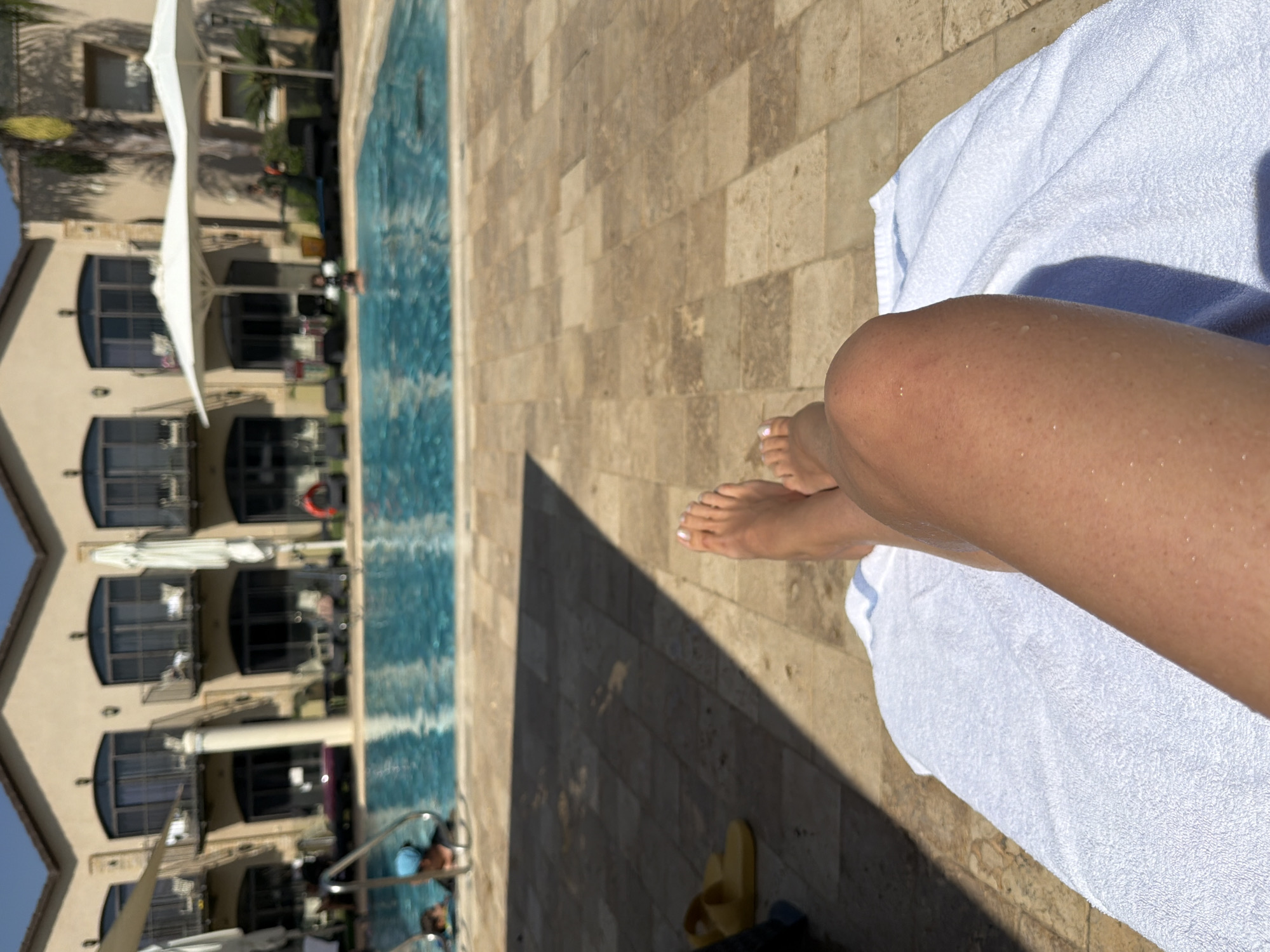 A first-person perspective shot of a person's legs and feet resting on a white towel at a resort swimming pool. The pool, with its clear blue water, is in the background, and there are a few people swimming in it. The resort building, with its neutral-colored facade and balconies, is also visible. Yellow flip-flops are placed on the tiled ground. The scene suggests a relaxing day at a pool in the Galil Elyon Regional Council, Israel.