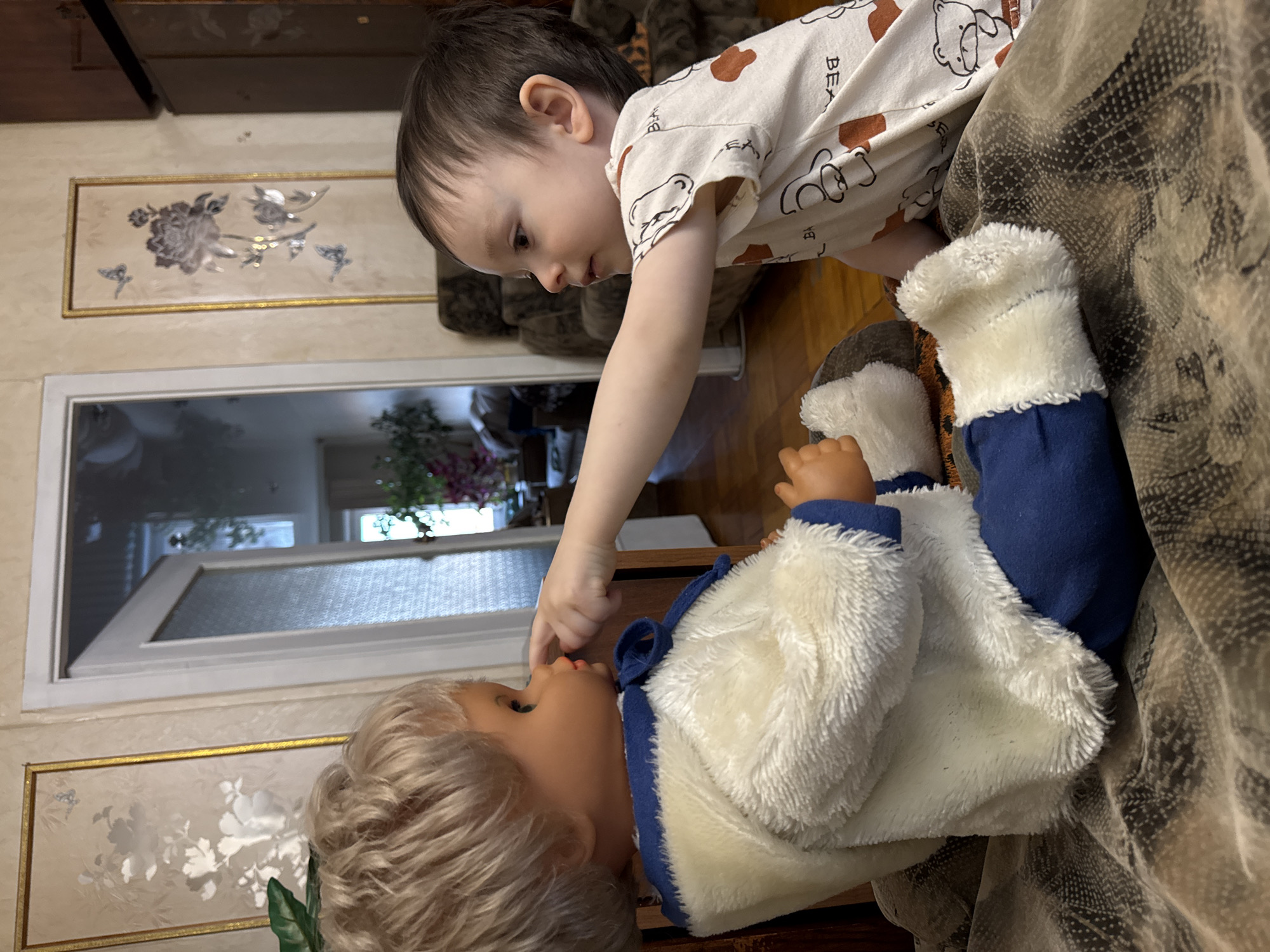 A young child with dark hair, wearing a white t-shirt featuring brown bear outlines and the word "BEAR," gently interacts with a doll. The child is leaning forward and down, extending their right arm to delicately touch the doll's nose or mouth with a finger. The doll has blonde hair, is dressed in a white fluffy jacket with blue trim, and blue pants, lying on its back on a patterned blanket or cushion. The child's expression appears focused and tender, suggesting a moment of curious or affectionate play.
The setting is indoors, characteristic of a home in Izhevsk, Russia. A light-colored wall serves as the background, adorned with a rectangular framed picture showcasing metallic or silver-toned floral patterns against a beige base, enclosed within a golden frame. Another similar frame is partially visible. A window with frosted or patterned glass is seen in the background, allowing natural light into the room, with a glimpse of greenery outside. Sections of dark wooden furniture are visible in the upper and lower portions of the frame. The time of day appears to be daytime due to the natural light.