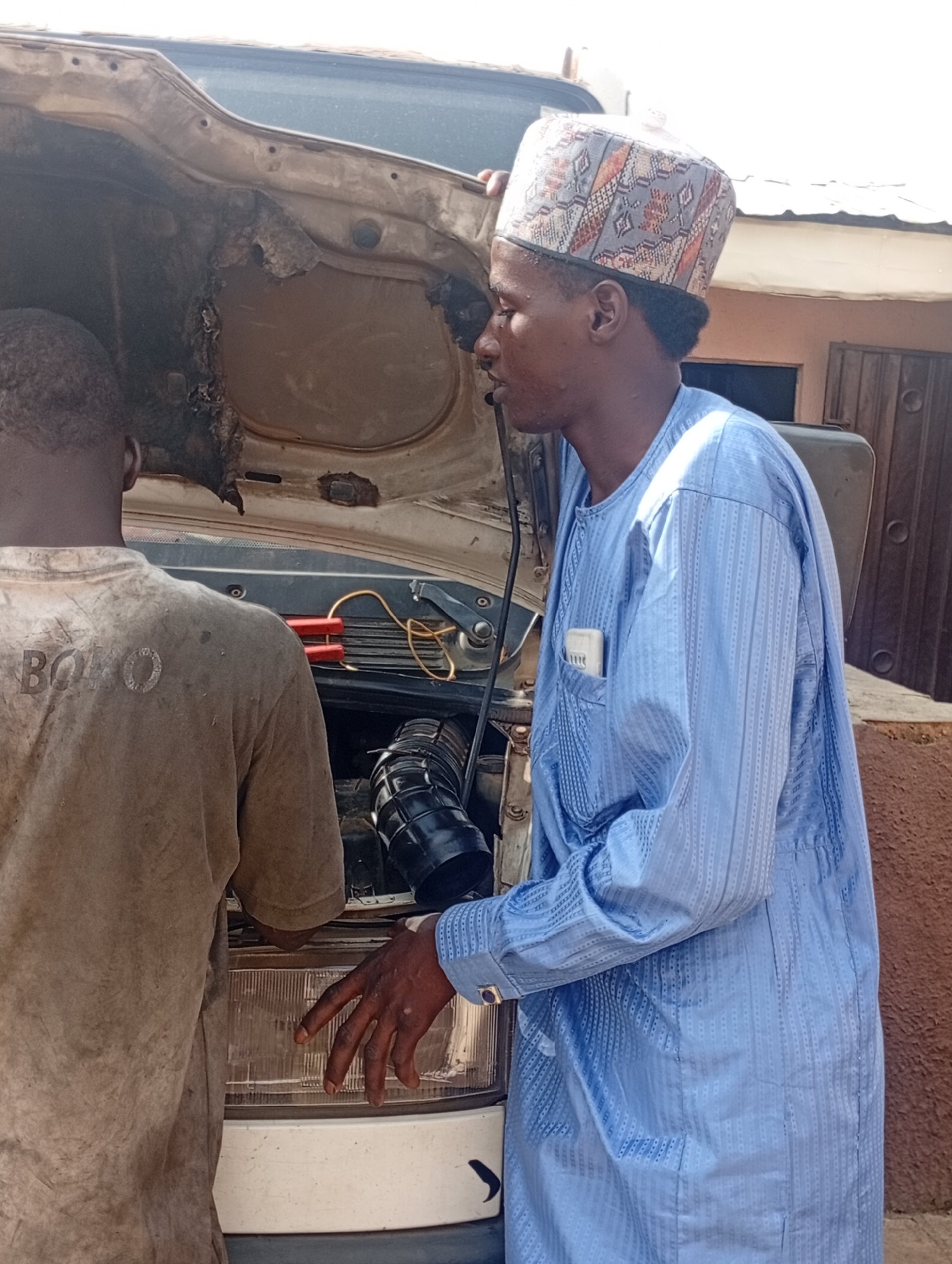 Two men are working on the open engine of a vehicle in an outdoor setting. One man, wearing a light brown, dirt-stained t-shirt with the word "BOKO" visible on the back, stands with his back to the camera, facing the engine. The other man, dressed in a blue patterned robe and a patterned cap, stands to the right, facing the engine and looking at it intently. He appears to be the focus of the image. His right hand is visible, touching the engine area. A small white object, possibly a pen, is tucked into the breast pocket of his robe.
The scene suggests a mechanical repair or inspection. The hood of the vehicle is propped open, revealing various engine components and wiring. There are jumper cables with red clamps visible connected to the engine. The background shows a building with wooden doors and windows, and a reddish-brown textured wall, consistent with an outdoor environment. The lighting suggests it is daytime, with bright sunlight casting shadows. The overall impression is one of everyday activity and work being done on the vehicle. There is no clear indication of the weather, but the bright sunlight suggests it is likely fair.