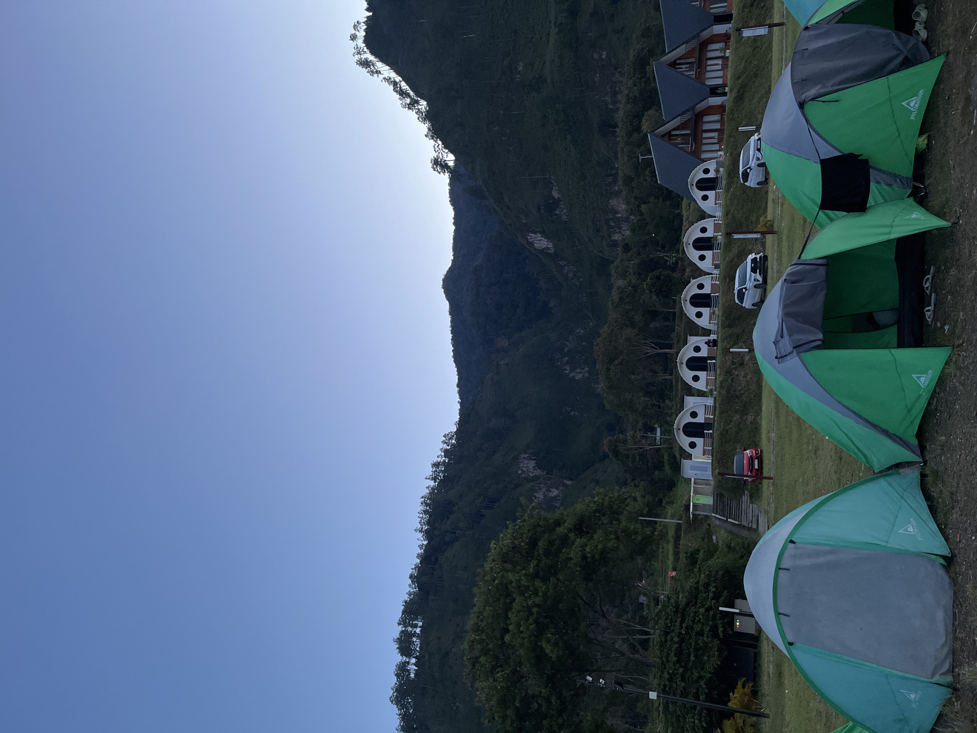 The image depicts an outdoor scene at a campsite in Merodot, Indonesia, likely during dawn or dusk due to the clear, pale blue sky and the subdued lighting. In the foreground, several tents are pitched on a grassy area. Two green and gray tents are most prominent, one open with its interior visible, and another closed. Other tents are partially visible to the left and right.
Behind the tents, a row of distinct dome-shaped structures, possibly cabins or unique camping accommodations, are visible. Further back, several modern wooden houses with dark roofs are situated on a higher level of the terrain. Several cars are parked in front of the houses and dome structures, including two white SUVs and a red sedan.
The background is dominated by a large, densely forested mountain range. The slopes are covered in trees, with some lighter rock formations visible on the cliff faces. The trees at the very top of the mountain range are silhouetted against the sky. The overall environment suggests a peaceful, natural setting, popular for outdoor recreation and lodging.