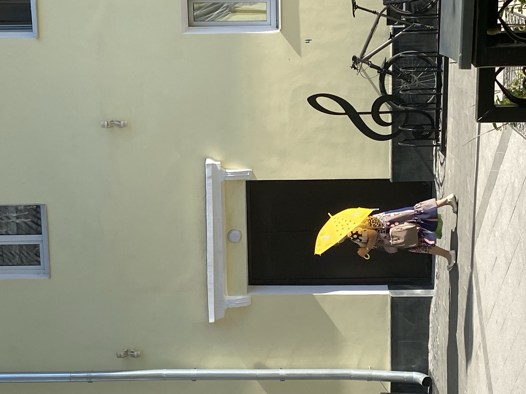 The image shows a street scene in what appears to be Заволжье, Russia. The scene is dominated by the side of a building with a light yellow facade. On the facade are two windows, a metal pipe running horizontally along the bottom, and a door with a white frame detail. A large metal treble clef is mounted on the wall, next to a bicycle rack with at least two bicycles parked. A person is walking on the sidewalk, holding a bright yellow umbrella. The person is dressed in a patterned dress and carrying a bag. The sidewalk is made of gray paving stones.