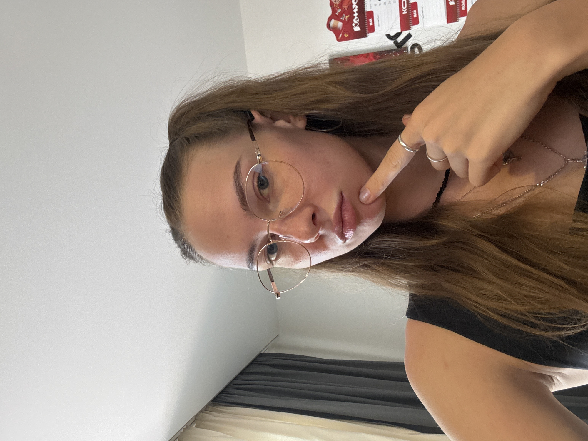A young woman with long brown hair pulled into a ponytail, wearing round, gold-rimmed glasses, a black tank top, and a choker necklace with a cross pendant, is pictured from the chest up. She is looking directly at the camera with her right index finger touching her chin. She is wearing two silver rings on her right hand. The background features a white ceiling, a curtain with vertical grey and cream stripes, and a calendar with red and white squares on the right side of the frame. The lighting suggests an indoor environment.