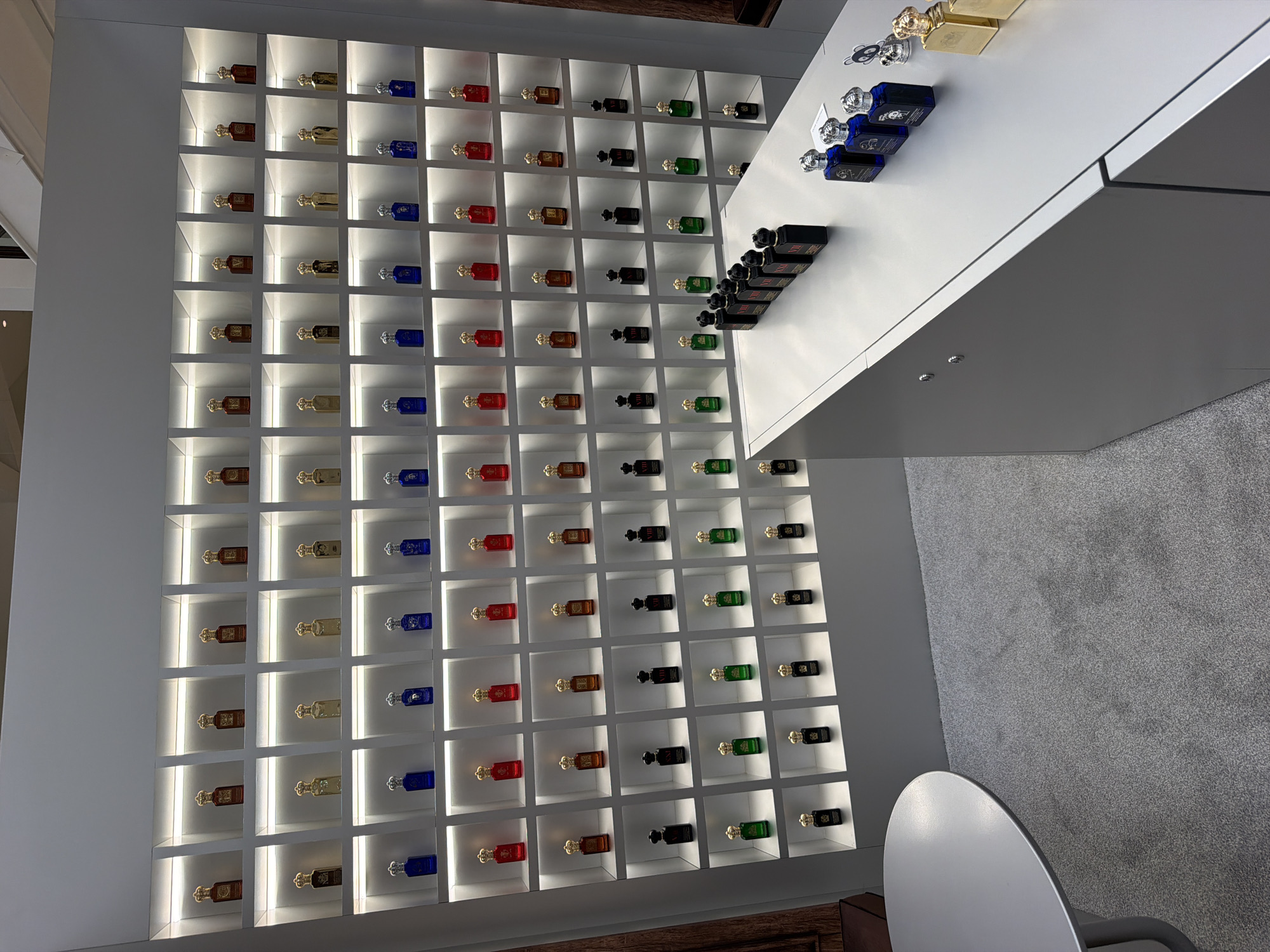 The image shows a retail display of perfumes in Aventura, Florida, United States of America. The display features a large wall unit with multiple illuminated shelves, each holding an array of perfume bottles. The bottles are arranged in rows and columns, with variations in color and cap design. To the right of the wall display is a modern, gray counter with more perfume bottles displayed on its surface. Some bottles are dark, while others are a vibrant blue. There are also a few decorative items, including what appears to be a small silver skull-shaped ornament and gold-colored perfume bottles. A round, light gray table is visible on the left side of the image, placed on a light gray carpeted floor. The overall environment suggests a clean and organized retail space, likely a department store or a dedicated perfume boutique. There are no people visible in the image.
