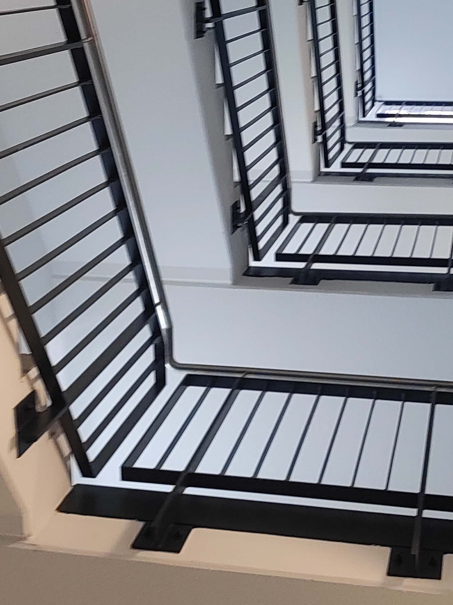 The image shows an abstract view looking up at a spiral staircase in an interior, likely a building. The staircase features black metal railings with a repeating diagonal pattern against white walls and steps. The perspective is from below, looking upwards, creating a sense of depth and geometric repetition. The lighting appears to be natural, coming from above, illuminating the white surfaces and casting shadows from the railings. The scene is clean and modern, suggesting an architectural structure. No people or specific locations in Fürth, Germany are visible or identifiable.