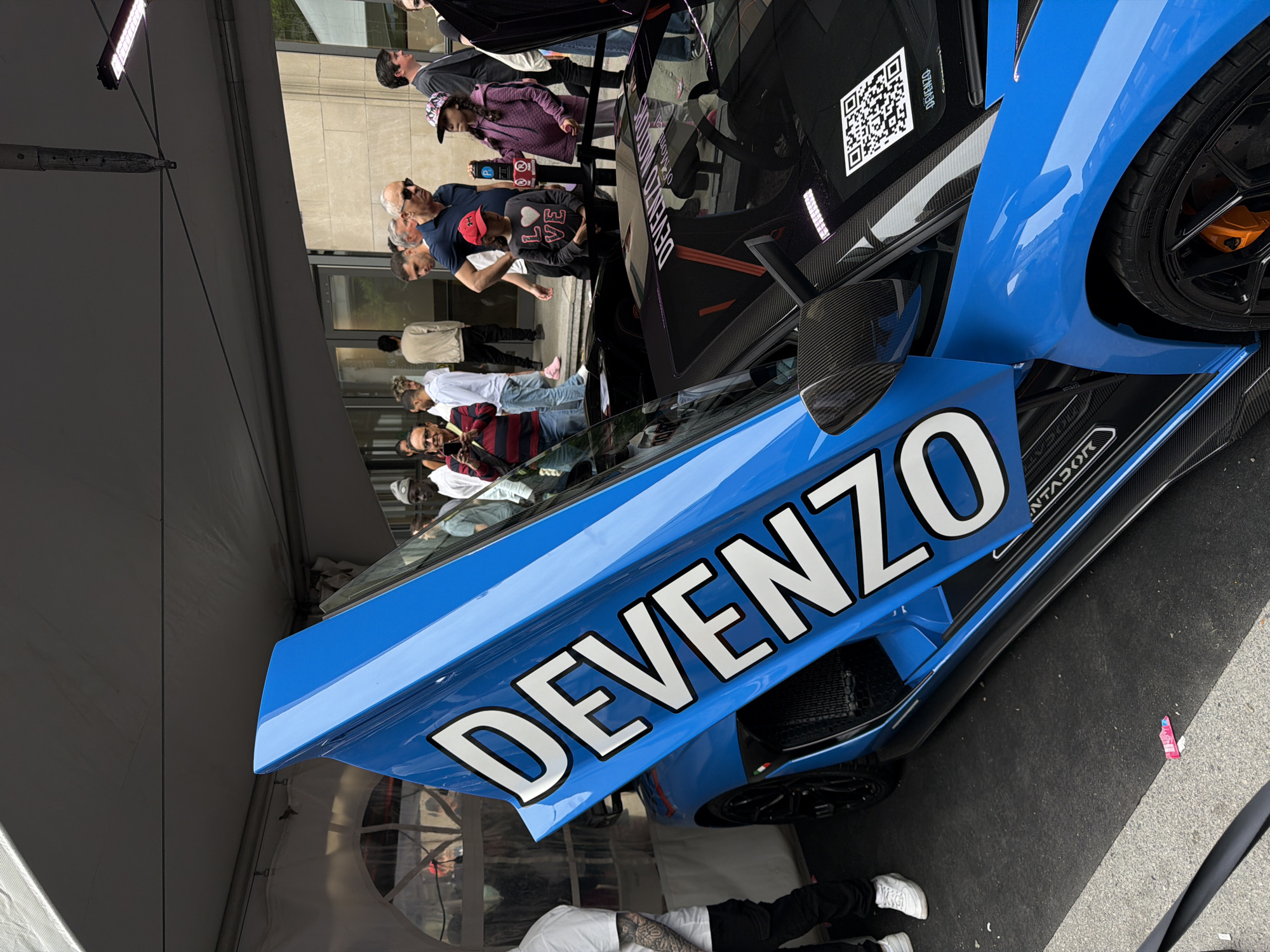 The image depicts a blue sports car with the word "DEVENZO" prominently displayed on its side. The car is surrounded by people, suggesting it's on display or at an event. The scene is outdoors, potentially on a street or in a designated area for showcasing vehicles. Details like a QR code on the car's window, carbon fiber accents, and high-performance tires are visible. The presence of a tent-like structure indicates that the event is held under cover or protection from the elements. However, I can't determine if the picture was taken in the city of Montreal, Canada. More clues would be needed to establish a definitive link to that location.