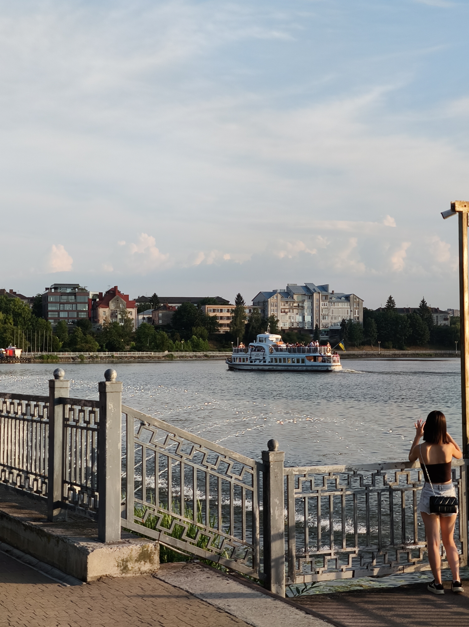 The image depicts a scenic view of Ternopil Lake in Ternopil, Ukraine. In the foreground, a young woman with dark hair stands on a paved walkway, leaning on a decorative metal railing and looking out at the water. She is wearing a black tank top, denim shorts, and sneakers, and has a quilted handbag slung over her shoulder. Her hands are raised, and she appears to be taking a photo or signaling with her fingers.
The lake is calm, with a white passenger boat, filled with people, sailing across its surface. The boat has a Ukrainian flag flying from its stern. In the background, a picturesque cityscape lines the shore, featuring a mix of modern and older buildings amidst lush green trees. The sky is a pale blue with scattered white clouds, suggesting it is late afternoon or early evening. A lamppost is visible to the right of the frame. The scene conveys a sense of leisure and natural beauty within an urban setting.