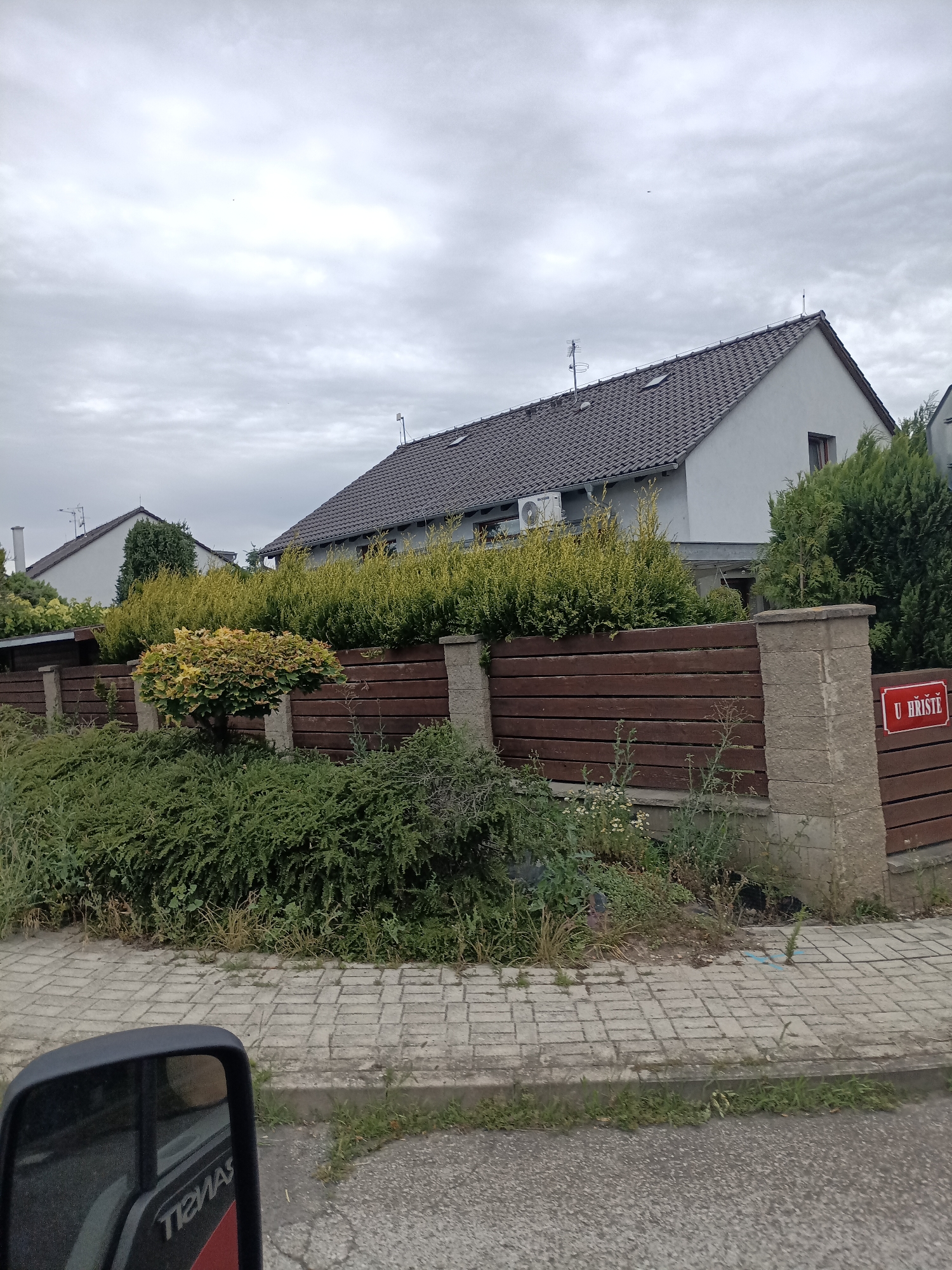 This is a photograph taken from a vehicle, showing a residential area on a cloudy day. In the foreground, the side mirror of the vehicle is visible, reflecting a portion of the vehicle's interior and a red and white logo with the text "TIEMAS" in reverse.
Beyond the vehicle, a paved sidewalk runs alongside a wooden fence. The fence is constructed with horizontal dark brown wooden planks supported by concrete pillars. A small, ornamental tree with yellow and green foliage stands in front of the fence, surrounded by lush green bushes. Behind the fence, two houses are visible. The house on the right has a dark grey tiled roof and a light grey exterior wall. An outdoor air conditioning unit is mounted on its wall. The house on the left also has a grey tiled roof and a white exterior.
Attached to one of the concrete fence pillars is a red sign with white text that reads "U HŘIŠTĚ". This translates to "At the playground" in Czech, suggesting the location of the property. The overall atmosphere is overcast and still, with no people visible in the frame. The scene depicts a quiet suburban or semi-rural setting in Svémyslice, Czechia, likely during the daytime.