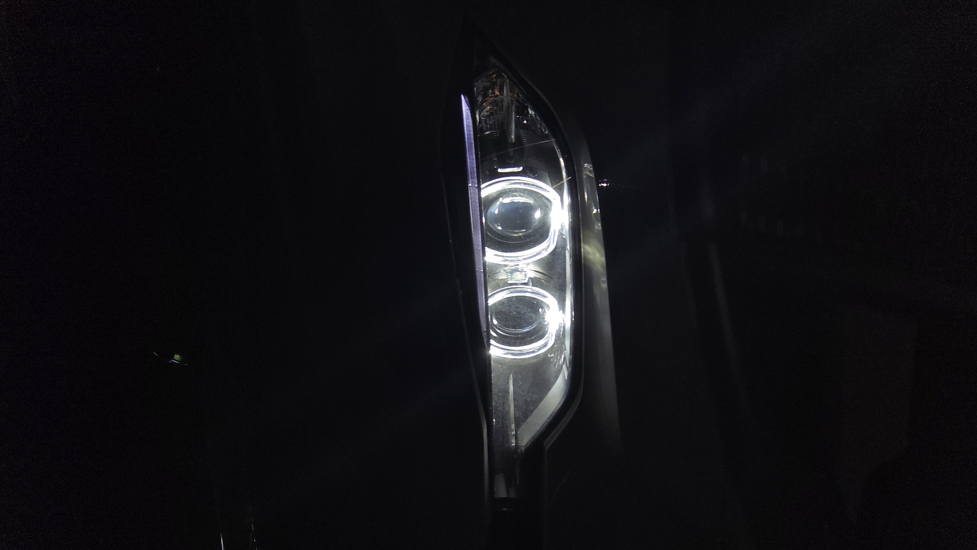 The media file displays a close-up of an illuminated car headlight, set against a dark, featureless background, strongly suggesting nighttime or very low-light conditions. The headlight, positioned vertically within the frame, is the sole prominent subject. It features two bright, circular projector lenses stacked one above the other, emitting a cool white light. To the left of these main lenses, a distinct vertical strip of white LED lights is visible, likely functioning as a daytime running light or position light.
The surrounding area is almost entirely black, with only faint reflections visible on the dark surfaces immediately adjacent to the headlight, possibly indicating the car's bodywork. The image focuses exclusively on this automotive detail, highlighting its design and illumination. There are no human subjects, discernible activity, or specific weather conditions depicted. No visible text appears in the image. This static shot captures an automotive feature in Sofia, Bulgaria, emphasizing its artificial light against the natural darkness.