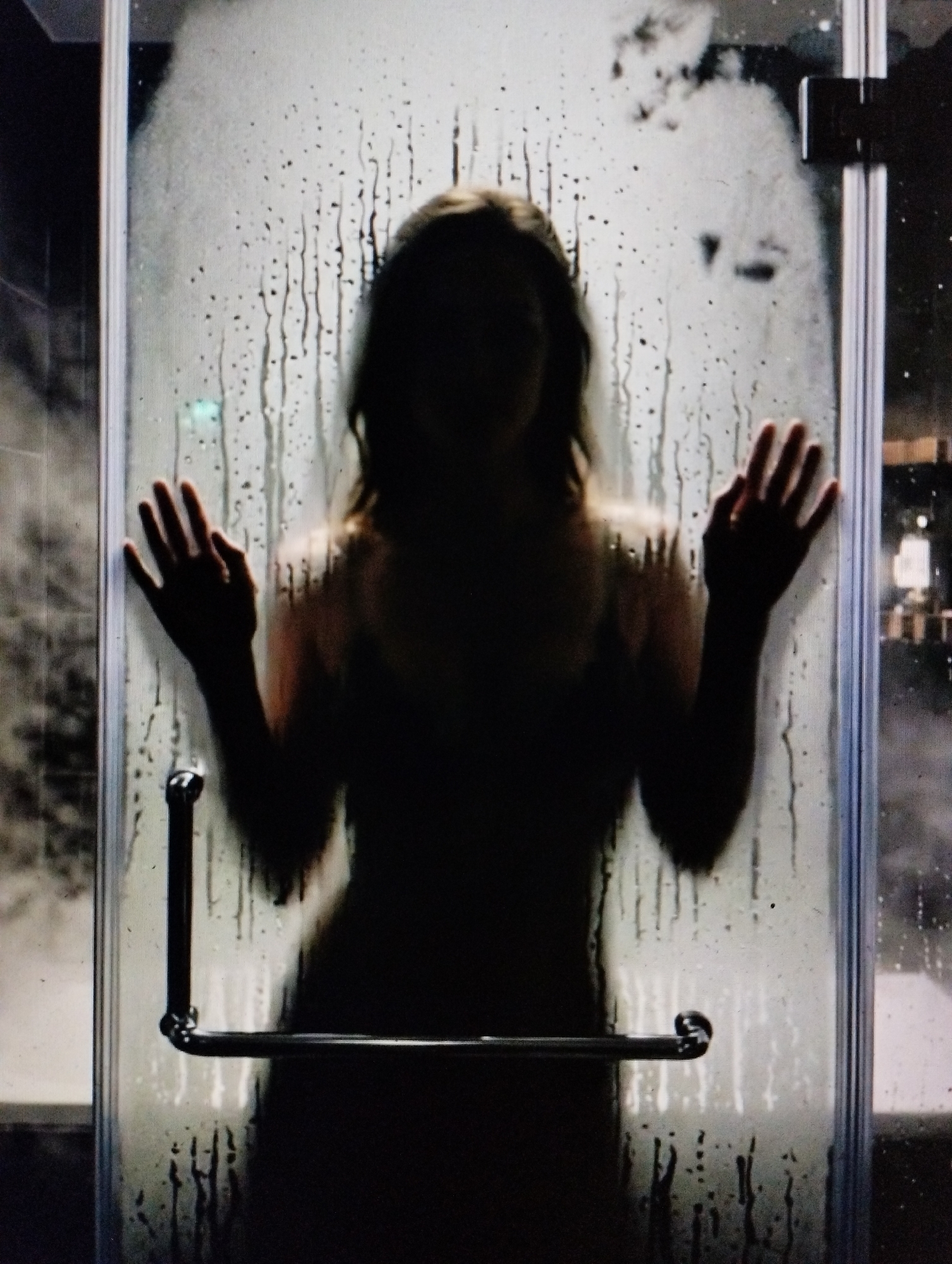 A person is silhouetted behind a steamy shower door, their hands pressed against the glass. Water droplets and streaks obscure the view of their body, rendering them as a dark, indistinct form. A metal shower door handle is visible on the lower part of the frame. The background is dark and out of focus, with faint lights suggesting an urban setting at night.