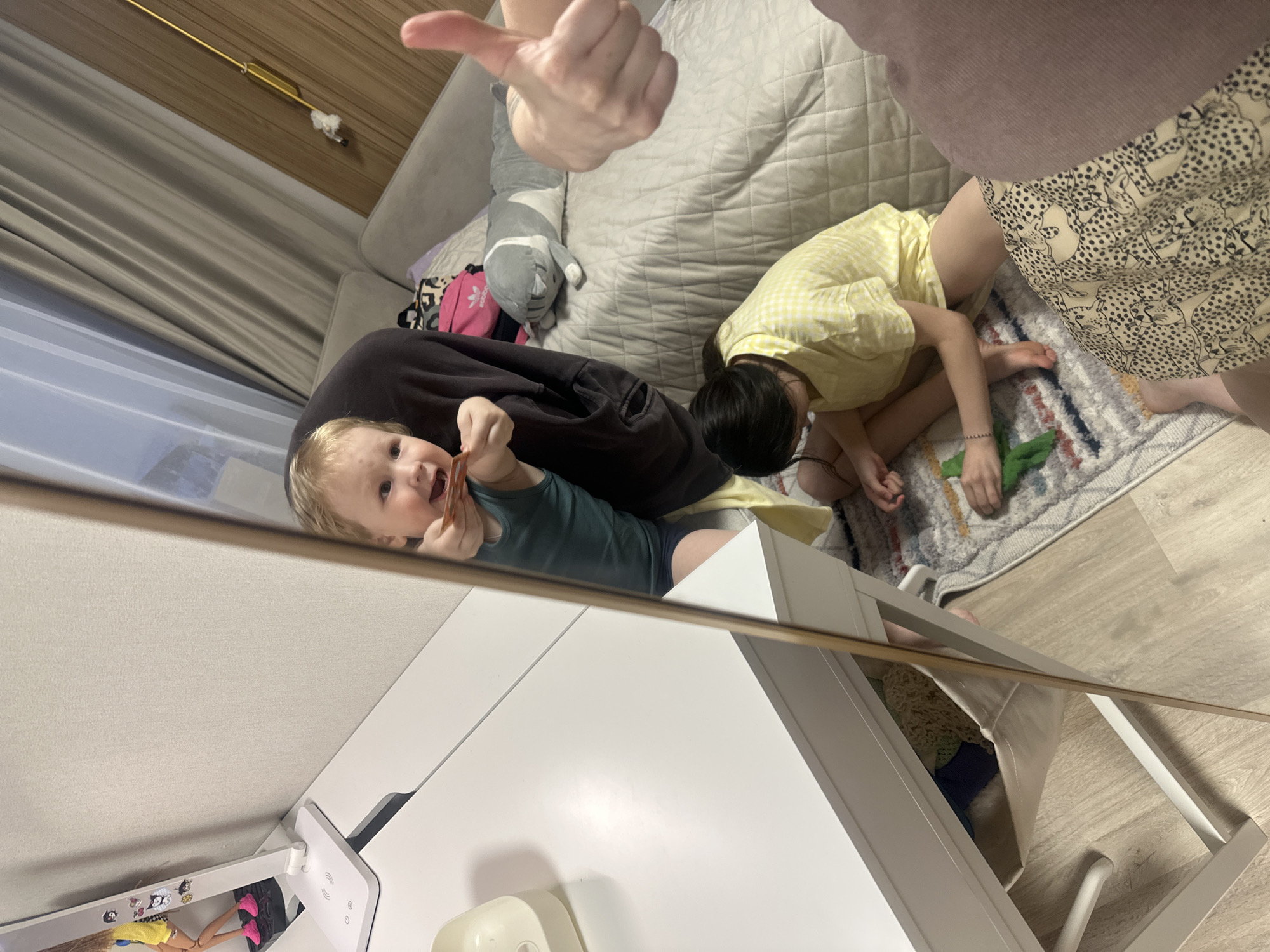 A mirrored view of a room in Votkinsk, Russia reveals two children and one adult. The first child, a toddler with blonde hair and fair skin, is reflected in the mirror. He is wearing a green shirt and dark shorts, and he is holding a small object to his mouth. In the background, a second child with black hair, wearing a yellow dress, is kneeling on a rug on the floor. The rug has muted shades of grey, brown and green. An adult wearing patterned shorts and a purple shirt is also in the frame. The adult's hand is raised with thumb extended up. The setting includes a grey couch, a white desk, a toy-filled white shelf, and a few small soft toys on the couch.