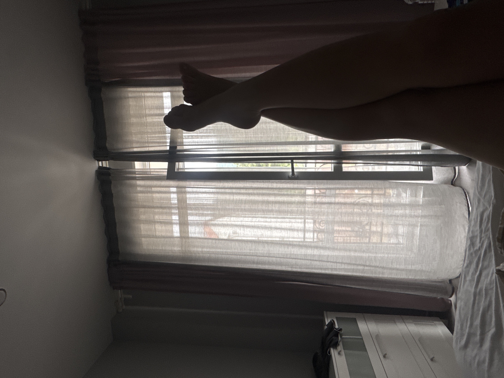 The image shows a pair of crossed legs and feet silhouetted against a window. The window has light-colored curtains. Beyond the window, there is a glimpse of an urban scene, potentially showing architectural elements. To the side of the window, there is a white storage cabinet or dresser. The legs and feet are the primary subjects. The lighting is dim, which contributes to the silhouetted effect. There is no evidence in the image to suggest the location is Gelendzhik, Russia.