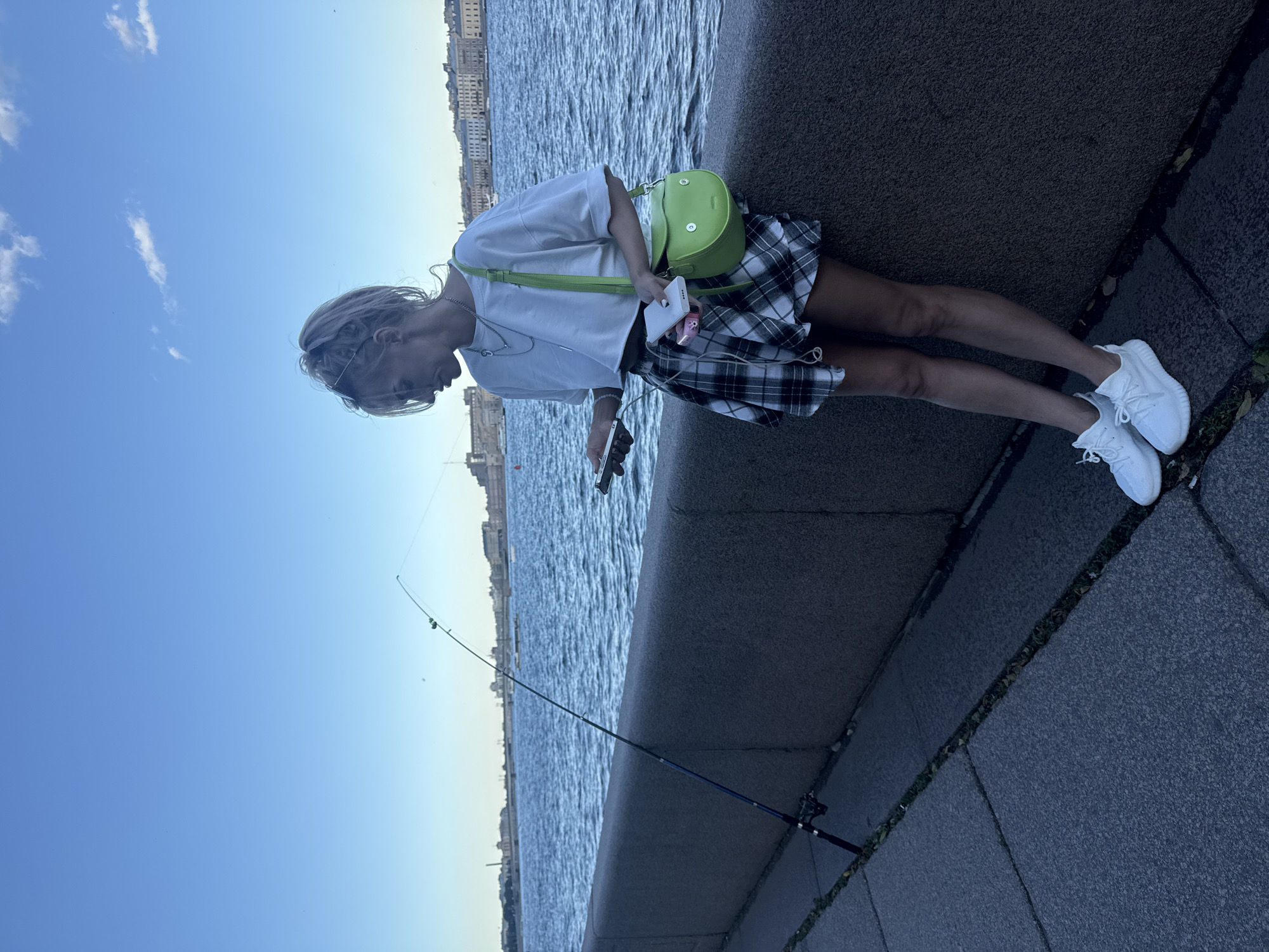 A young woman with blonde hair tied in a ponytail stands leaning against a concrete embankment next to a body of water, likely the Neva River in Saint Petersburg, Russia. She is dressed in a white crop top, a green and white plaid skirt, and white sneakers. A bright green, rounded crossbody bag with a playful face design hangs from her shoulder. In her hands, she holds a smartphone and what appears to be a portable charger. To her left, a fishing rod is propped up on the embankment. The background shows a cityscape with buildings and a clear blue sky.