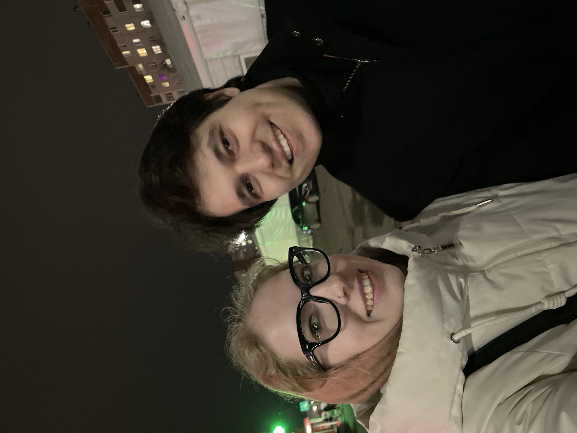 Two people, one wearing black clothing and one wearing a white jacket and eyeglasses, pose for a selfie. In the background are buildings and nighttime street illumination. The city is Izhevsk, Russia.