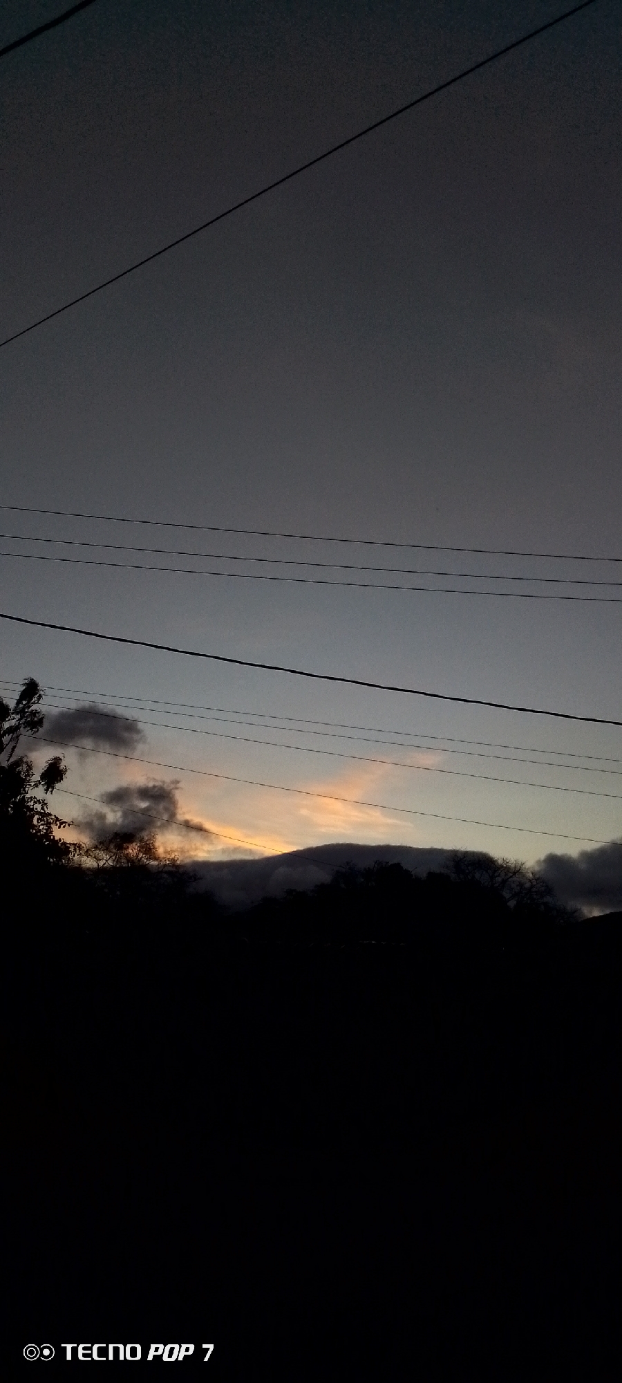 The image presents a serene twilight landscape, likely captured at dusk or early night, in Altagracia, Venezuela. The sky transitions from a dark grey-blue at the top to a softer band of light orange and yellow hues near the horizon, indicative of a fading sunset. Several dark utility lines horizontally traverse the upper and middle sections of the frame, with one thicker line running diagonally across the top right.
Along the horizon, the silhouettes of a mountain range or hills are prominently featured, occupying the lower third of the image. Dark, wispy clouds are interspersed among these silhouettes and in the mid-ground, some illuminated by the last vestiges of sunlight. On the far left, the dark outline of trees or dense foliage is visible against the muted sky.
The overall scene conveys a sense of peaceful transition. In the bottom-left corner, a watermark reads "TECNO POP 7" accompanied by a camera icon, identifying the camera used to take the picture.