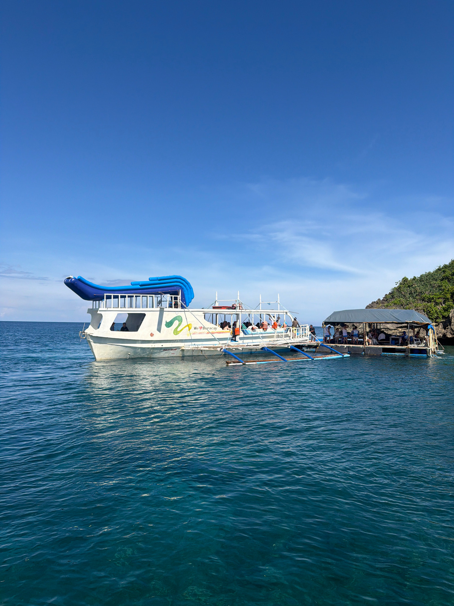 In the city of Nabas, Philippines, a clear blue sky stretches over a tranquil turquoise sea. Moored near a rocky, tree-covered island are two vessels. The primary vessel is a white, multi-deck boat with blue inflatable slides on its upper deck and the text "We <3 Boracay" on its side. This boat is occupied by several people. Alongside it, a second, smaller vessel, which appears to be a floating platform covered by a dark roof, is also carrying a group of passengers. The water around the boats is calm, with sunlight reflecting off its surface. The scene suggests a leisure activity, likely a tour or excursion in this picturesque location.