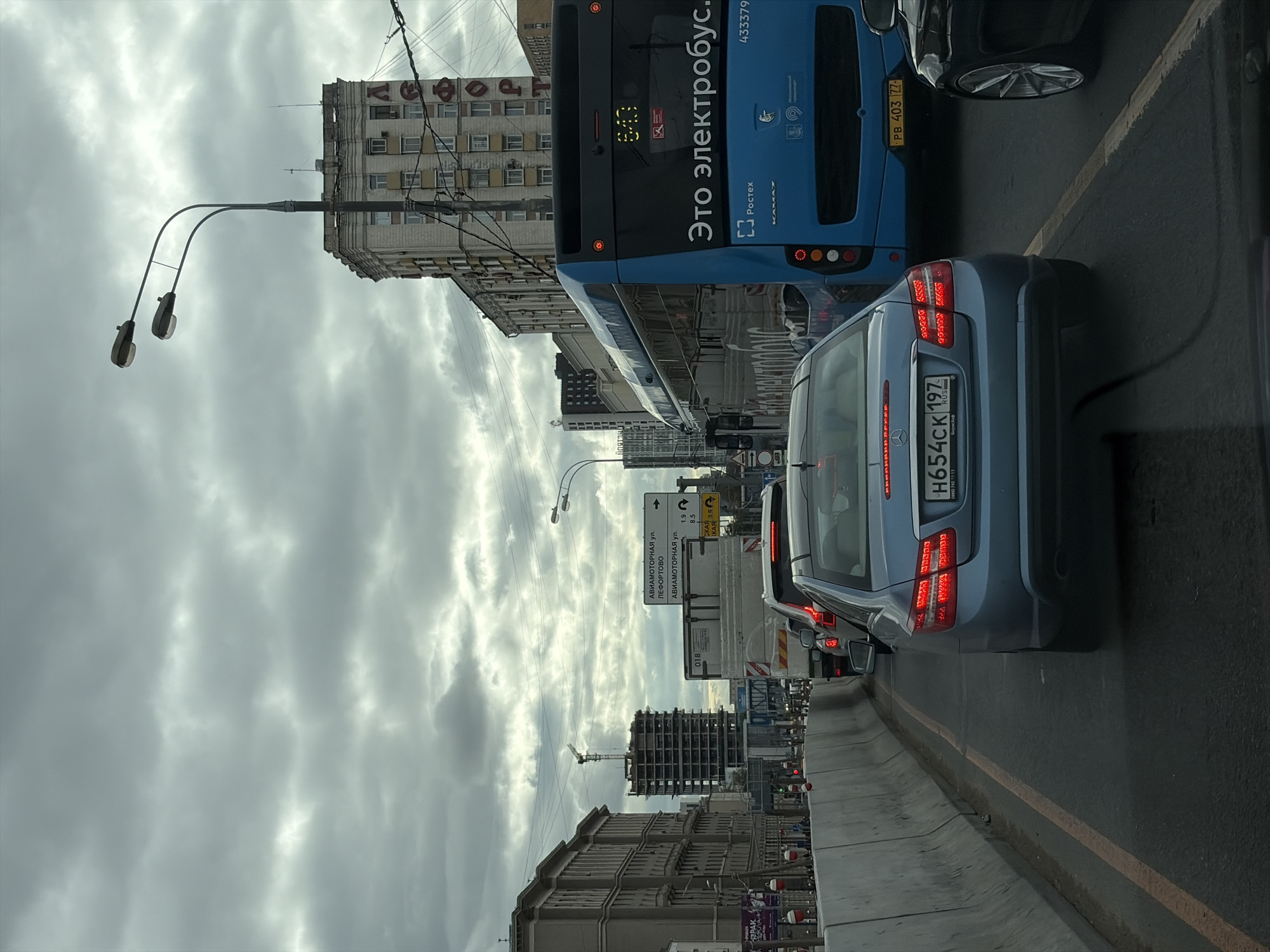 A traffic jam is captured on a cloudy day in Moscow, Russia. The viewpoint is from inside a vehicle. In the foreground, a light blue Mercedes-Benz sedan is visible with its brake lights illuminated. To its right, a black car's rear wheel and side skirt are seen. Further ahead and to the left, a concrete barrier lines the road, with some construction barriers and reflective markers. Beyond the barrier, construction is underway on a tall building with a crane. In the midground, a bright blue electric bus with "Это электробус." written on its side is prominent. Behind the bus, a white truck is stopped in traffic. Traffic signs are visible, one indicating "АВИАМОТОРНАЯ ул." with distances of 1.9 km and 8.5 km. Several multi-story buildings line the street, some appearing older with distinct architectural features, while others are modern high-rises under construction or completed. The sky is overcast with heavy gray clouds. The overall scene depicts a typical urban traffic congestion.