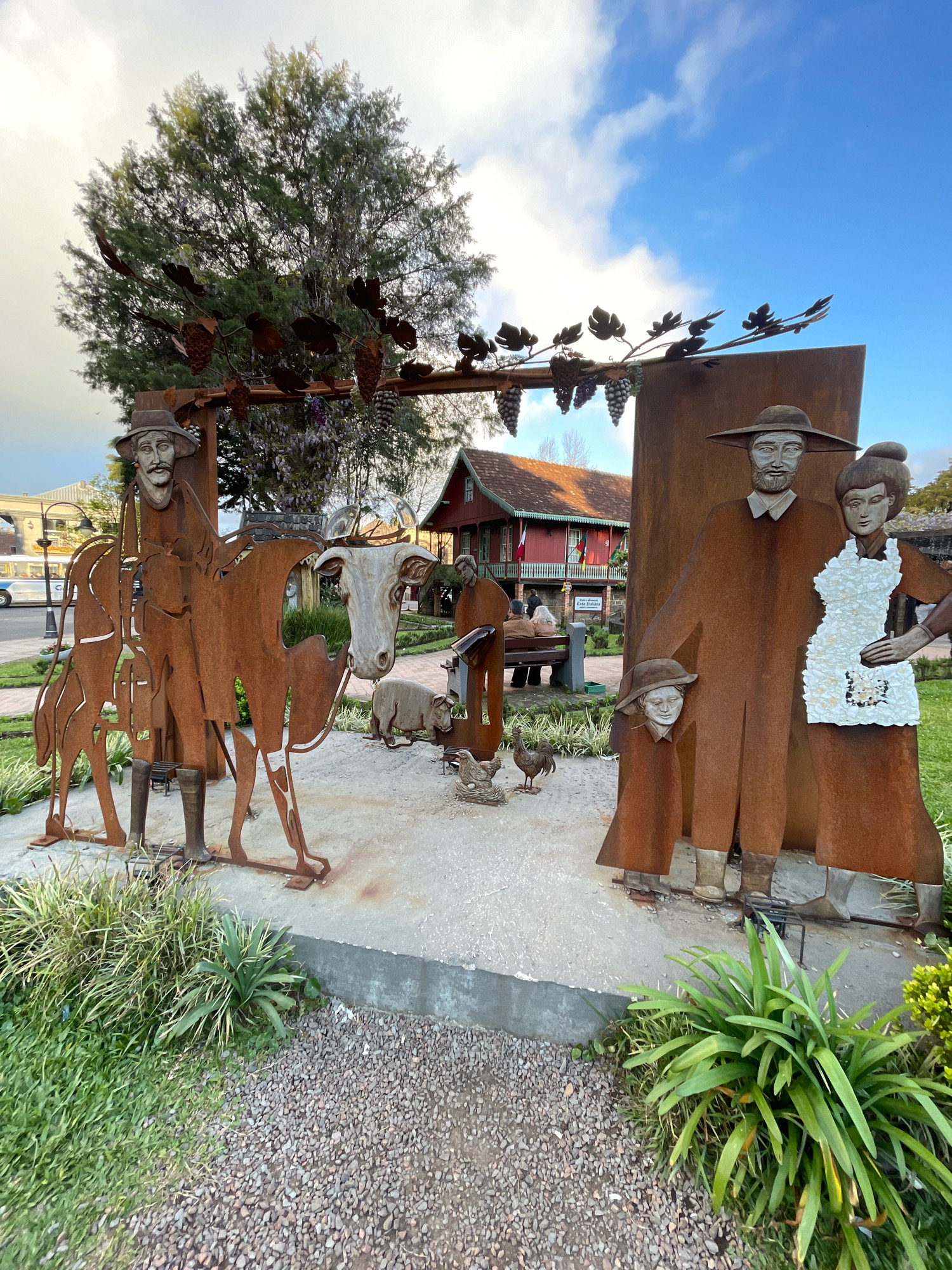 This image captures an outdoor art installation in Gramado, Brazil, featuring rustic metal sculptures that evoke a rural, possibly agricultural, theme. Dominating the foreground are several large, corten steel figures. To the left, a man on horseback, his face rendered with detail, is depicted with a stern expression. The horse and rider are stylized, with the metal cutouts suggesting the forms. In the center, a sculpture of a cow with a realistic, painted head stands next to a sheep. Further to the right, a family group is represented: a man, a woman wearing a white apron, and a child wearing a hat, all with detailed facial features and wearing broad-brimmed hats and cloaks. Small chicken sculptures are also scattered around the base of these main figures.
Behind the sculptures, a traditional-style building with red siding and white trim is visible, suggesting a European-influenced architectural style common in Gramado. A street with a white bus and other vehicles can be seen to the far left, indicating the installation is situated in a public area, likely a park or a plaza. The environment includes lush greenery with ornamental plants and gravel pathways. The sky is a vibrant blue with scattered white clouds, and a large tree with visible branches and foliage forms a backdrop to the scene. The overall impression is one of a community space celebrating heritage through art.