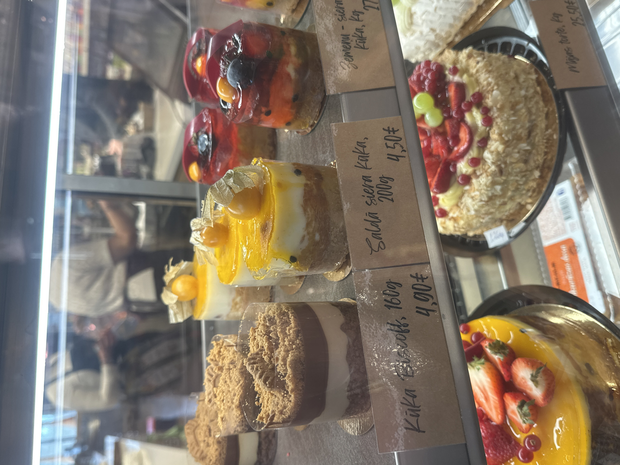 The image captures a display case filled with an assortment of cakes in Krustiņi, Latvia. The cakes vary in style and ingredients. In the top row, there are fruit topped cakes with a translucent glaze, a "Majas Torte" covered in crumbs and decorated with strawberries and grapes. The next row has slices of "Saldā siera kaka" cakes with golden topping. A lower row includes "Kūka Biscoff" dessert in clear cylindrical cups. Each cake has a small card displaying its name and price in euros. The background shows the reflection of a person and the interior of the shop.