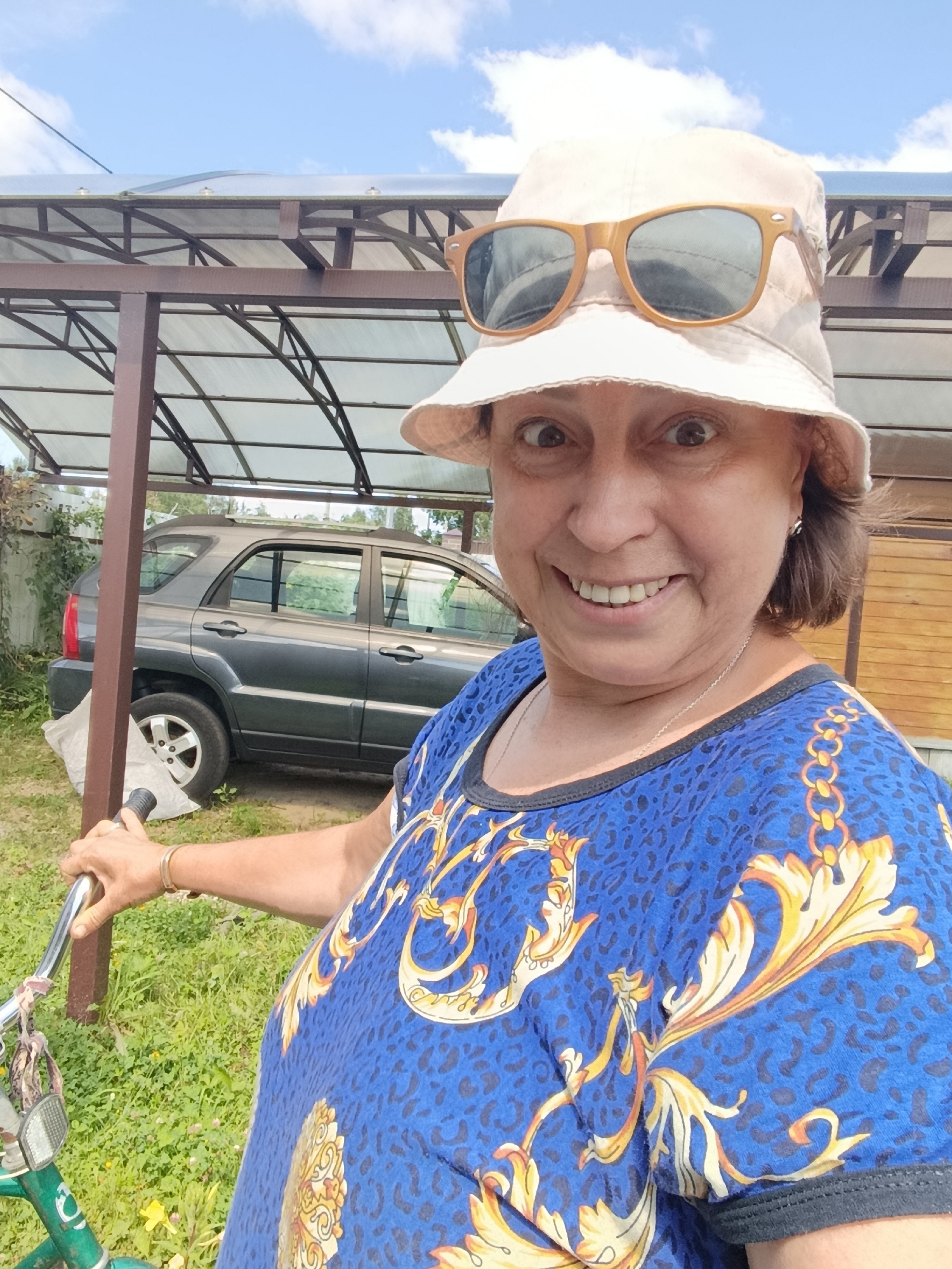 A middle-aged Caucasian woman wearing a beige bucket hat and sunglasses is taking a selfie outdoors. She has a wide smile and her eyes are wide open, looking at the camera. She is wearing a blue t-shirt with a gold and orange ornate pattern and leopard print accents. She is holding the handlebars of a green bicycle. In the background, a dark gray SUV is parked under a covered structure with a translucent roof and brown metal supports. The ground is covered in grass and some small yellow flowers. The sky is blue with scattered white clouds. The scene appears to be in a rural or suburban area, possibly a backyard or a dacha (summer house) in the city of Vereya, Russia.