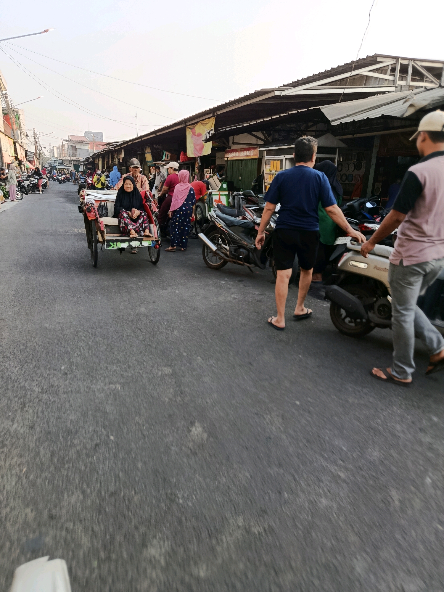 In the city of Bekasi, Indonesia, a street market scene unfolds under a hazy sky. The road is paved with asphalt, and on the right side, a row of market stalls with corrugated metal roofs are visible. Several motorbikes are parked along the side of the road in front of these stalls. Two men are walking away from the camera, one in a blue t-shirt and black shorts, the other in a pink and black shirt with grey pants. Both are wearing sandals. A light-colored scooter is parked on the right. On the left side of the road, a woman in a black hijab and patterned pants sits in a cycle rickshaw, which is moving forward. Other people are walking in the background, and more motorbikes are visible. The overall impression is a busy street market with pedestrian and vehicle traffic.