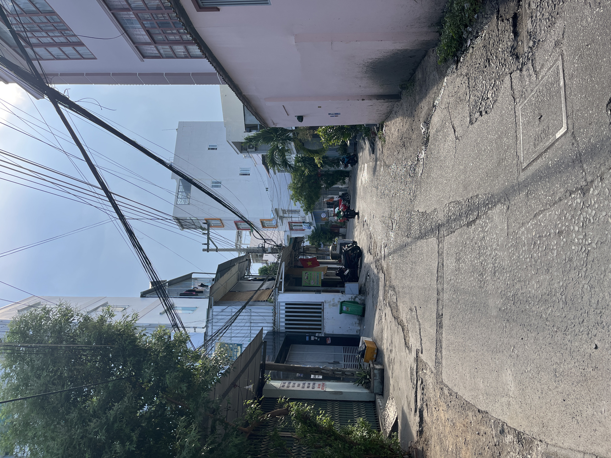 The image captures a narrow alleyway in Bac Nha Trang Ward, Vietnam. The alley is paved with cracked concrete, showing signs of wear and age. Tall, closely packed buildings line both sides of the alley, constructed with a mix of architectural styles and materials. Some buildings are painted in light pastel colors, while others show bare concrete or faded paint. Electrical wires densely crisscross overhead, creating a complex web against the sky. In the distance, a few motorcycles are visible parked along the alley, suggesting local activity. Some trash bins and plastic bags are visible in front of the buildings. Vegetation, including small trees and shrubs, grows sporadically along the edges of the alley, adding a touch of greenery. The overall impression is of a typical urban environment in Vietnam, characterized by narrow spaces, dense construction, and a mix of residential and commercial activity.