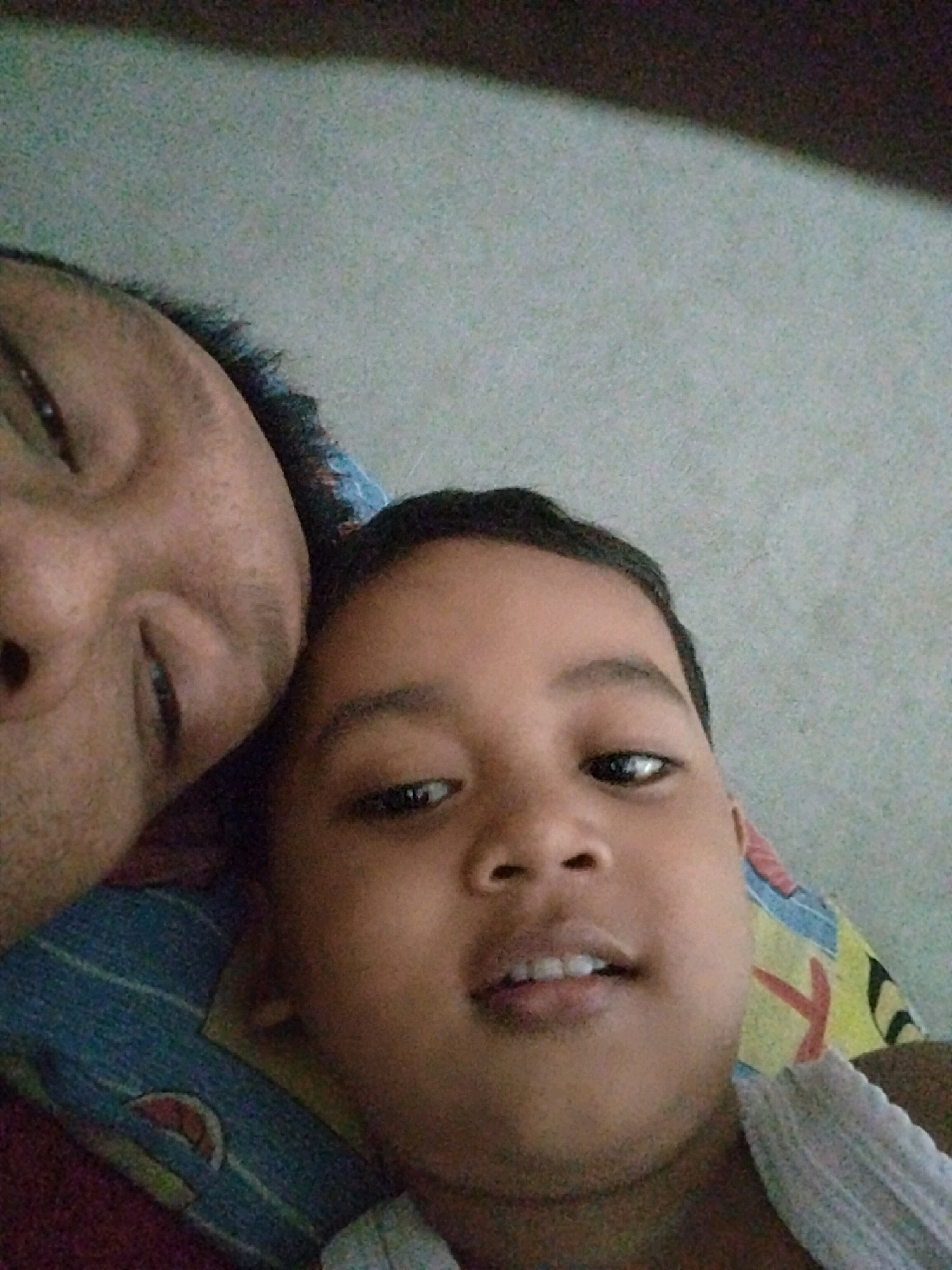 Two individuals are captured in a close-up selfie, with their faces filling most of the frame. The person on the left, positioned higher in the image, is an adult with a portion of their forehead, eye, and cheek visible. Their eye is open and looking towards the camera. The person on the right is a child, whose face is prominently displayed, looking directly at the camera with both eyes open. Their mouth is slightly open, revealing teeth. They are lying down, as indicated by the angle of their faces and the background. The background consists of a textured surface, possibly a wall, with a dark, diagonal line crossing the upper portion of the image. Below the subjects, a colorful patterned fabric is visible, appearing to be part of bedding. The image is taken indoors, likely in a residential setting, potentially in the city of Pepelegi, Indonesia, given the provided context. The lighting appears to be natural or from an overhead source, casting soft shadows.