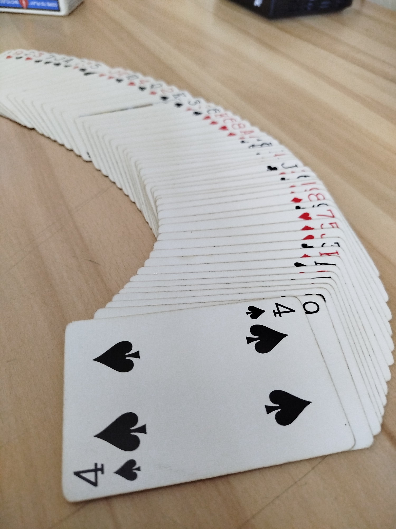 A fanned deck of playing cards lies on a wooden surface. The cards are white with black and red symbols. The leftmost cards are the most visible, showing the "Four of Spades" and the "King of Clubs". The other cards in the fan are partially visible, displaying various suits and numbers. The background is a plain wooden table. There are no people or animals in the image. The scene does not contain any elements that would indicate a specific location in Varna, Bulgaria.
