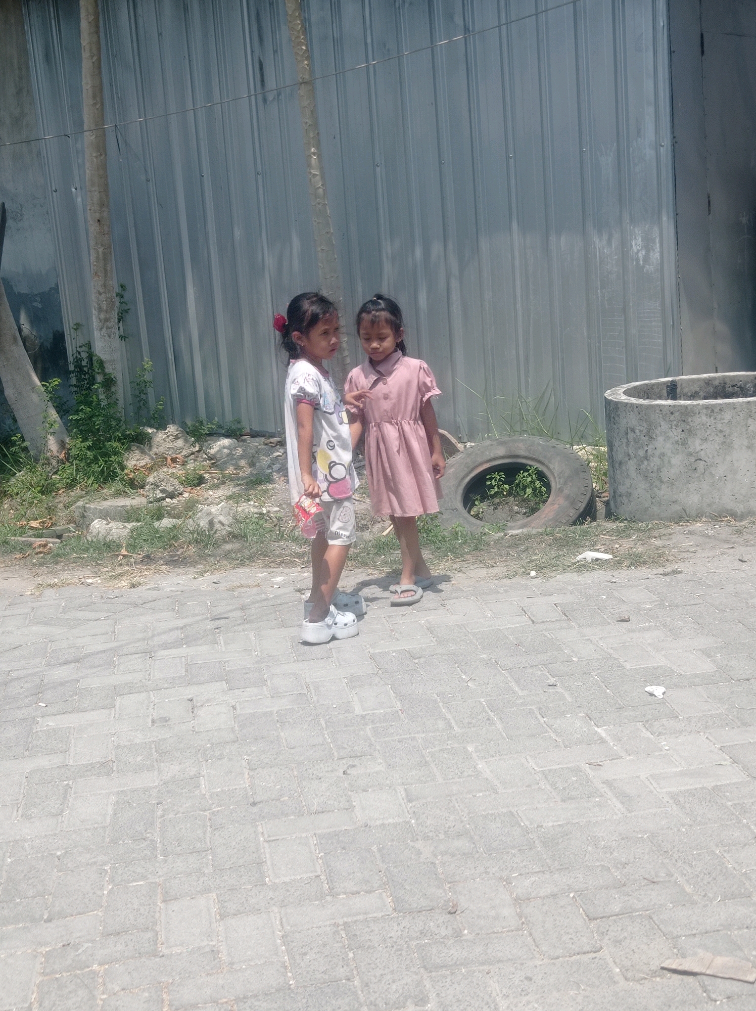Two young girls are standing on a paved pathway in front of a corrugated metal fence in Ngemplak, Indonesia. The girl on the left is wearing a white shirt with graphics and white platform clogs. She is holding a small, red and white package. The girl on the right is wearing a pink dress and gray flip-flops. They appear to be interacting with each other. To the right of the girls, there is a large concrete ring, and a deflated tire with a small plant growing inside it. The ground is a mixture of grass and dirt, and there is a thin, weathered tree trunk to the left of the girls. The scene is lit by bright sunlight, casting shadows on the ground.