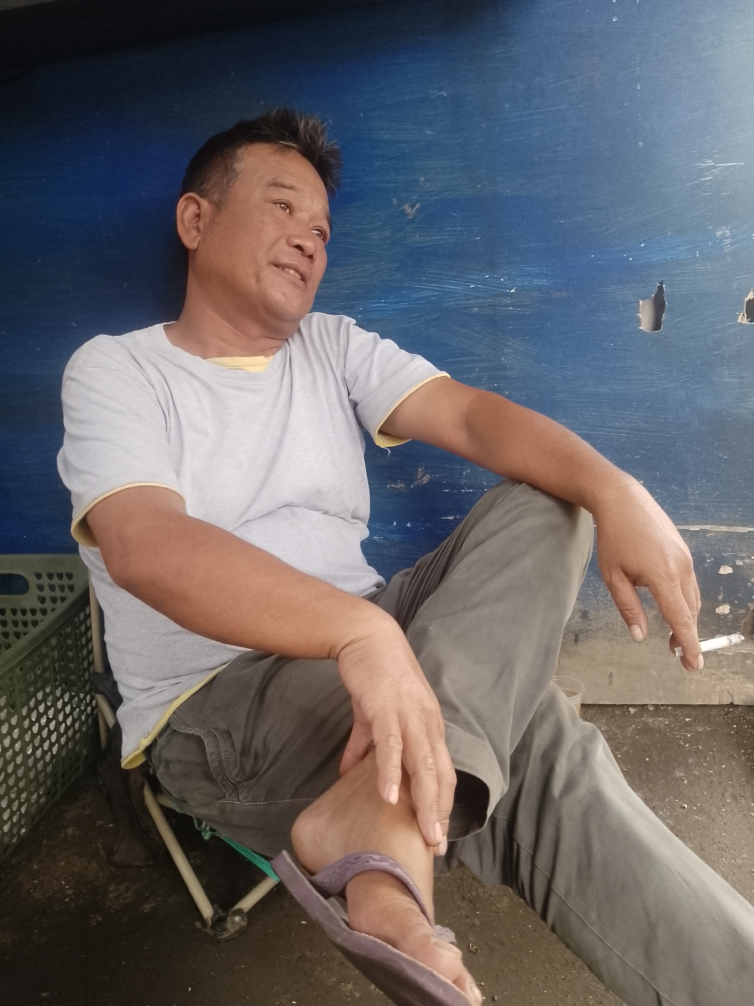 A medium shot captures a middle-aged Indonesian man in Cikalongkulon, Indonesia, sitting on a folding chair in front of a blue wall. He is wearing a light grey t-shirt with a yellow trim and grey pants. His right arm rests on his knee, and he is holding a lit cigarette between his index and middle fingers. His left hand is resting on his left thigh. His legs are crossed, with his left foot visible in a purple flip-flop. To the left of the man is a green plastic crate. The background consists of a textured blue wall with scuff marks and some small holes. The ground beneath the man appears to be dirt or concrete.