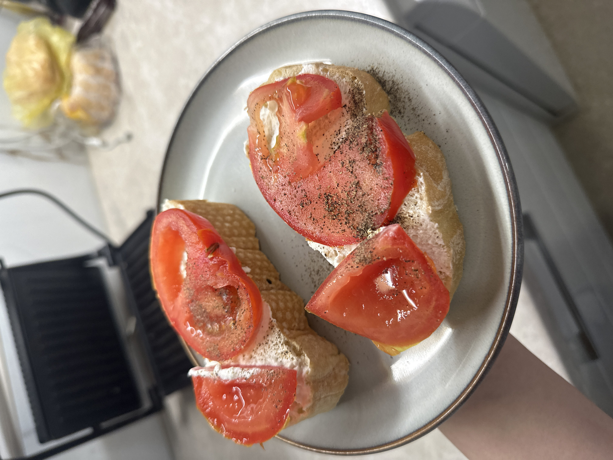 The image shows a close-up view of a plate with two pieces of bread topped with a white spread (possibly cream cheese), sliced tomatoes, and black pepper. The plate is light gray with a darker gray rim. The tomatoes appear fresh and juicy. The bread looks like a rustic type, possibly baguette slices. Part of a sandwich press is visible in the lower left corner. The background suggests an indoor setting with a light-colored countertop. The image is being held by a hand. There is no indication of the location being Budva, Montenegro, or any people.