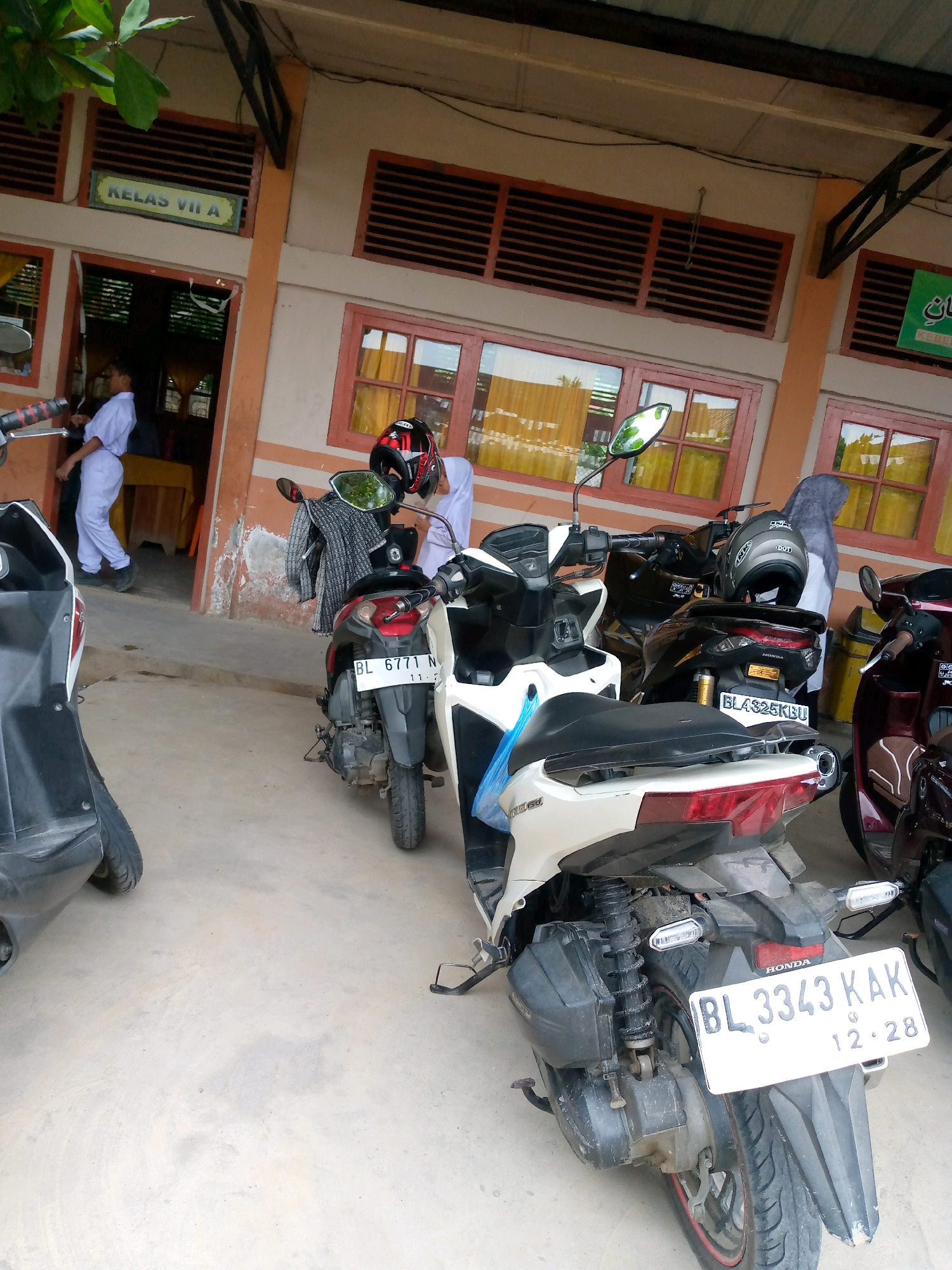 A group of motorcycles are parked outside a school building in the city of Mesjid, Indonesia. The building has an orange facade with multiple windows. A sign above a doorway reads "KELAS VII A" (Class VII A). Several students in uniform are seen near the entrance. One student is walking into the building, while others are near the parked motorcycles. The motorcycles are of various colors, including black, white, and maroon. Some of the motorcycles have helmets on their seats. The ground is a concrete surface. The weather appears to be overcast.