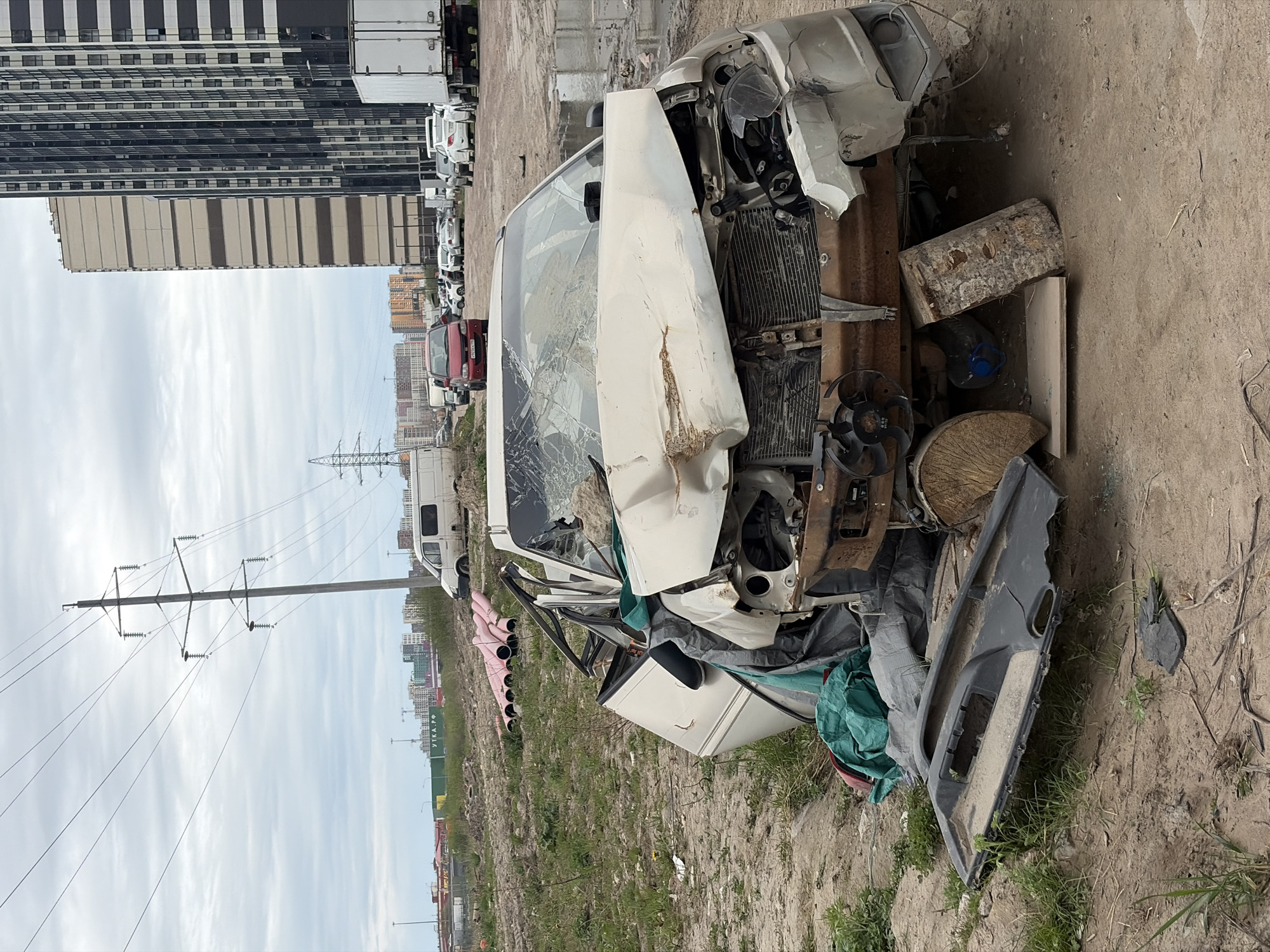 A wrecked white van is situated on a dirt and grass lot with a tall building and a row of parked cars and trucks in the background. The van is heavily damaged, with a shattered windshield, a crushed front end, and other parts scattered around it, including a detached door. A power line pole is visible in the distance. The setting appears to be a scrap yard or an abandoned area. Given the visible letters 'УТКА.РФ' on a green sign, and the architectural style of the buildings in the background, it is reasonable to assume that this location is in Russia. Based on the architectural style and the appearance of the landscape, it is plausible that this is somewhere on the outskirts of a large city, possibly in Saint Petersburg, Russia.